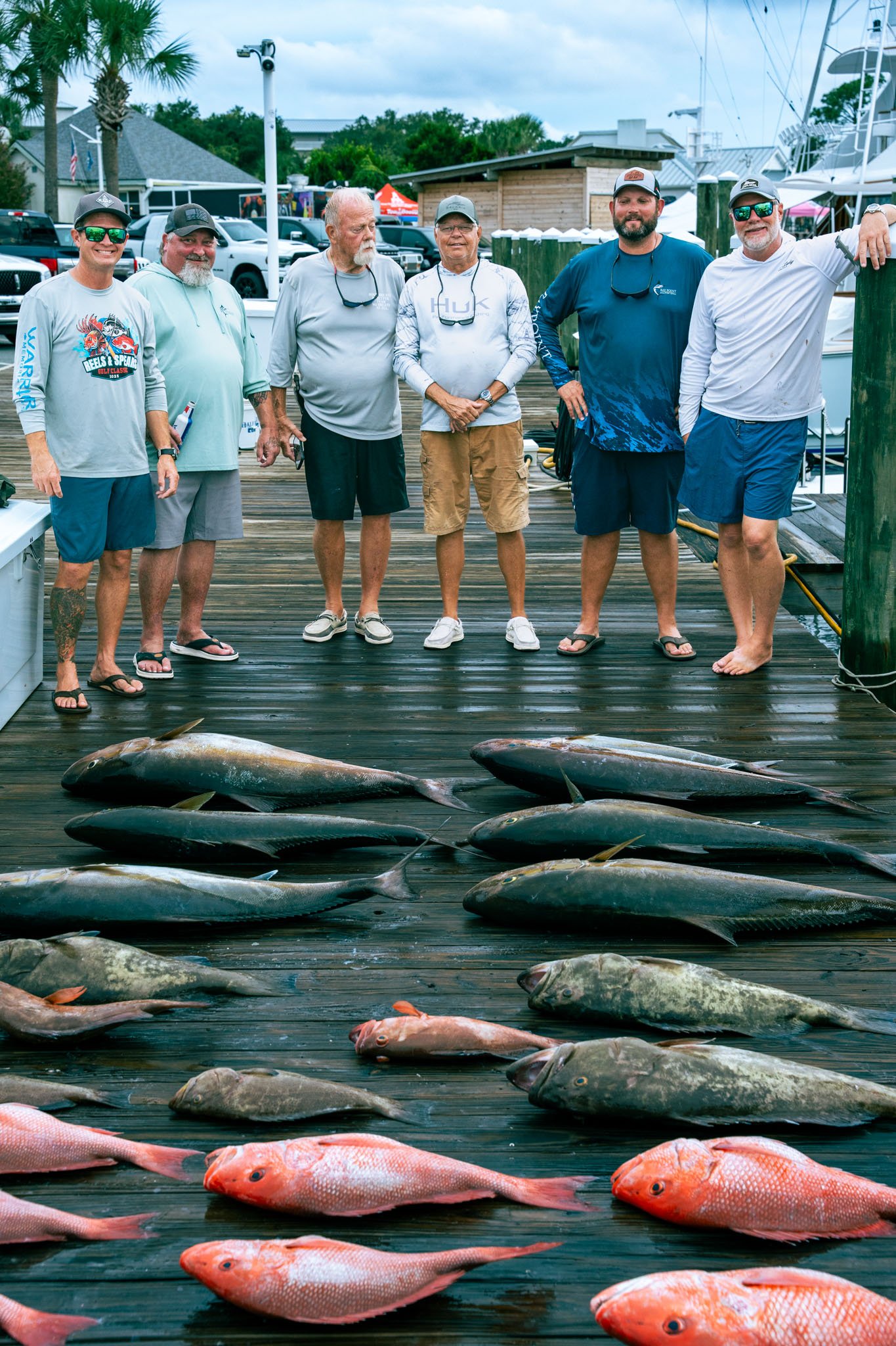 Group of seven men standing on a dock next to a boat, with fish laid out in front of them. Background includes parked cars, a building, and palm trees.