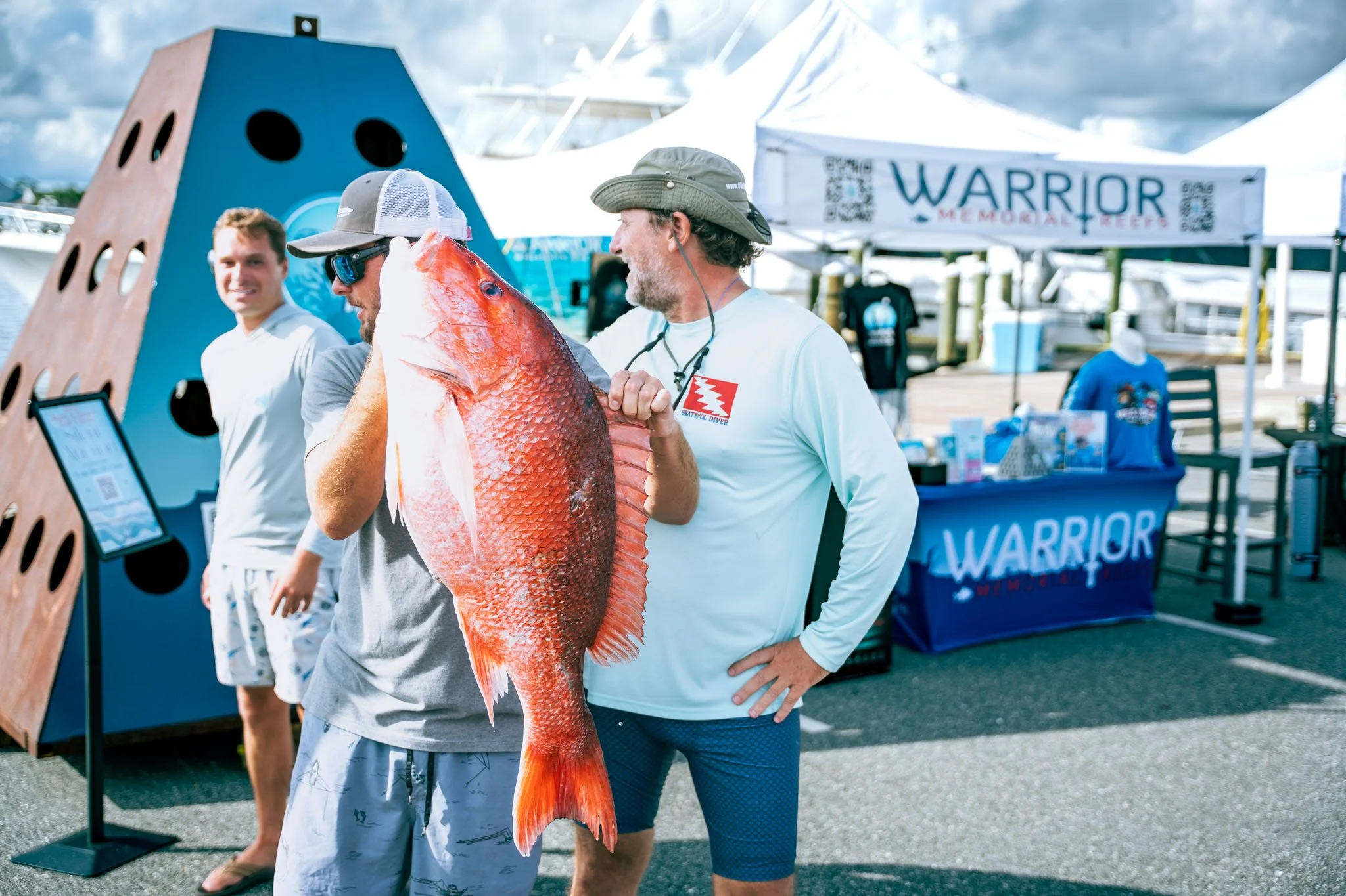 Two men holding a large red fish at Point South Marina - Bay Point (Panama City Beach) near boats and a tent with a 'Warrior Memorial Reefs' sign.