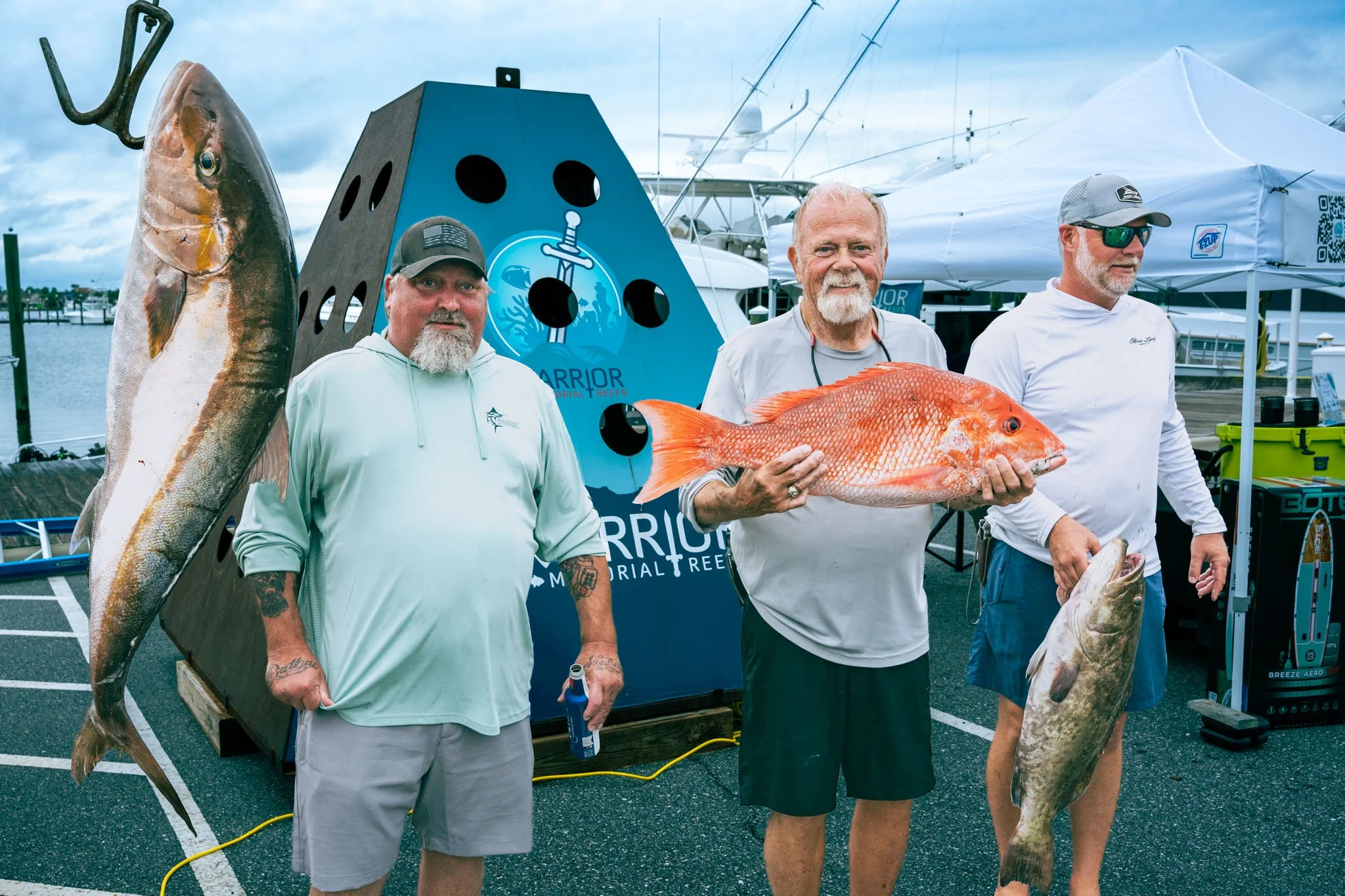 Three men standing in a marina holding large fish, with a display board and boats in the background.