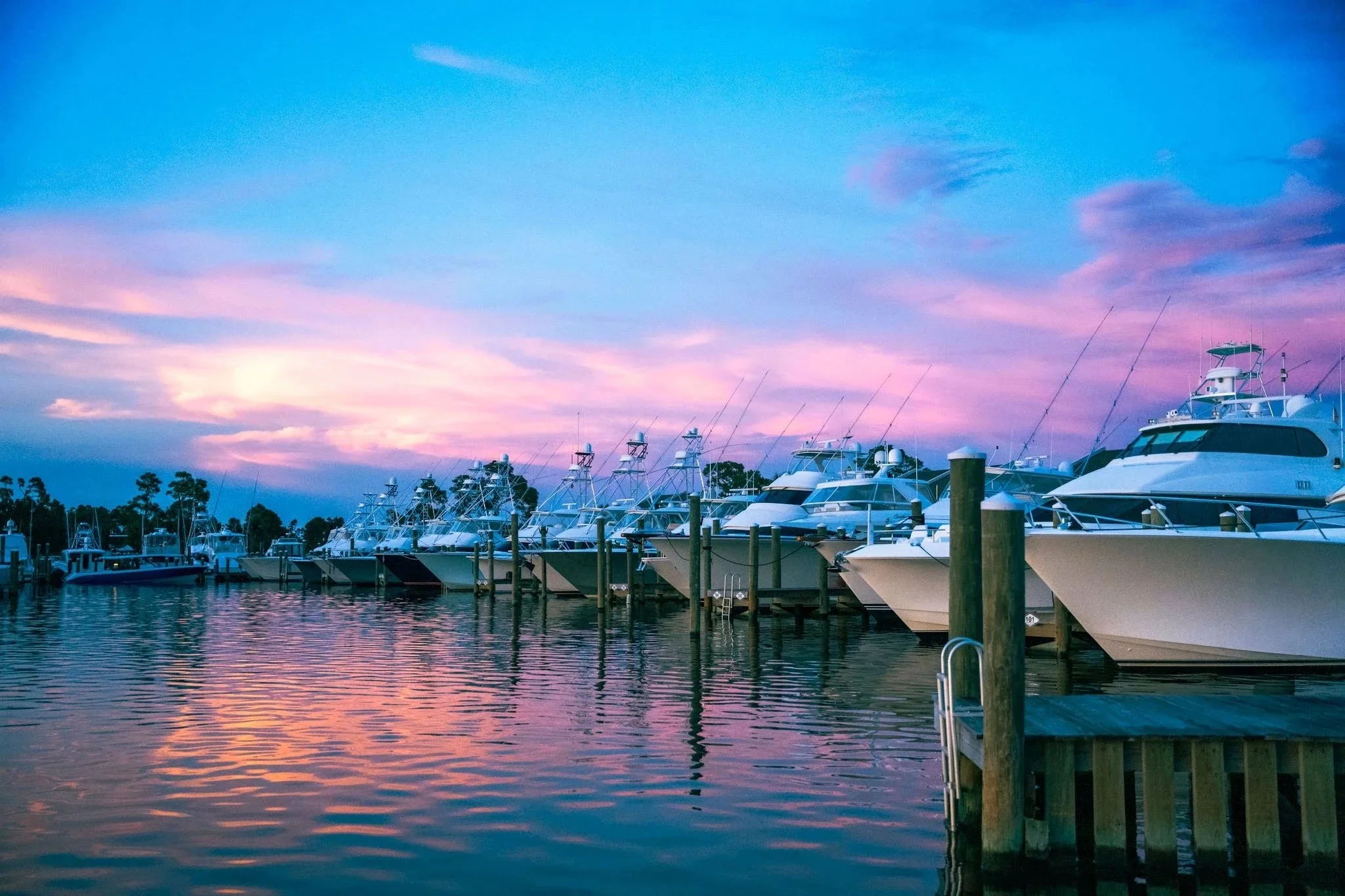 A marina with commercial fishing vessels/boats docked along Point South Marina - Bay Point (Panama City Beach) during sunset; sky painted in shades of pink and blue, calm water reflecting the colorful sky.