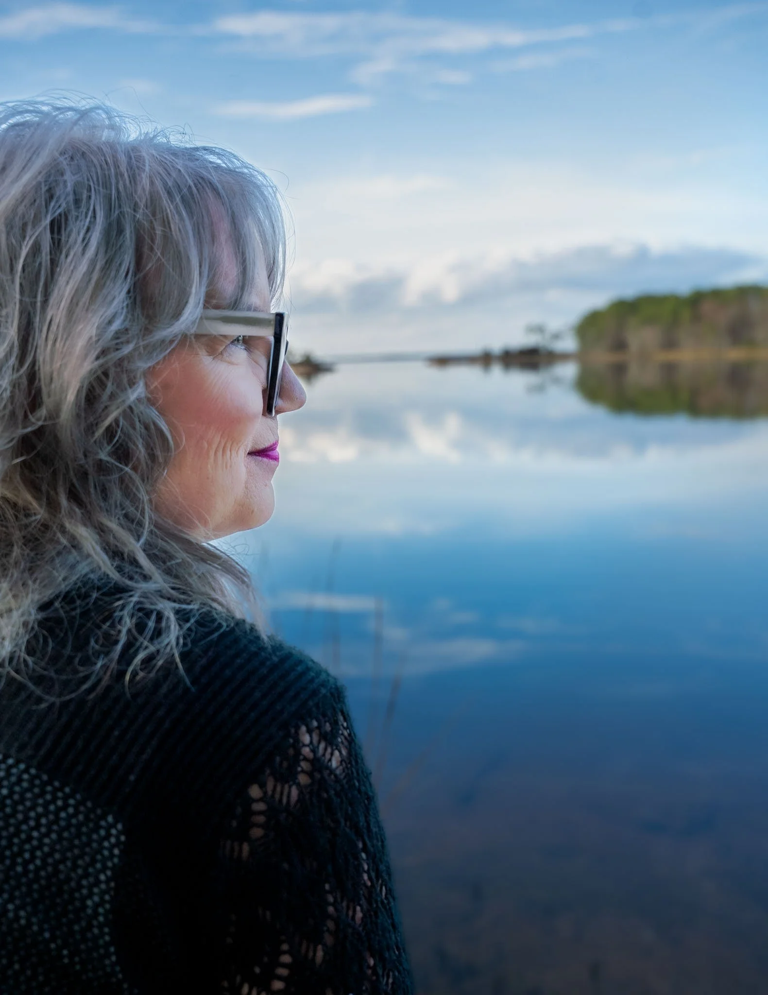 Side profile of an older woman pensively overlooking a lake/ estuarine area.