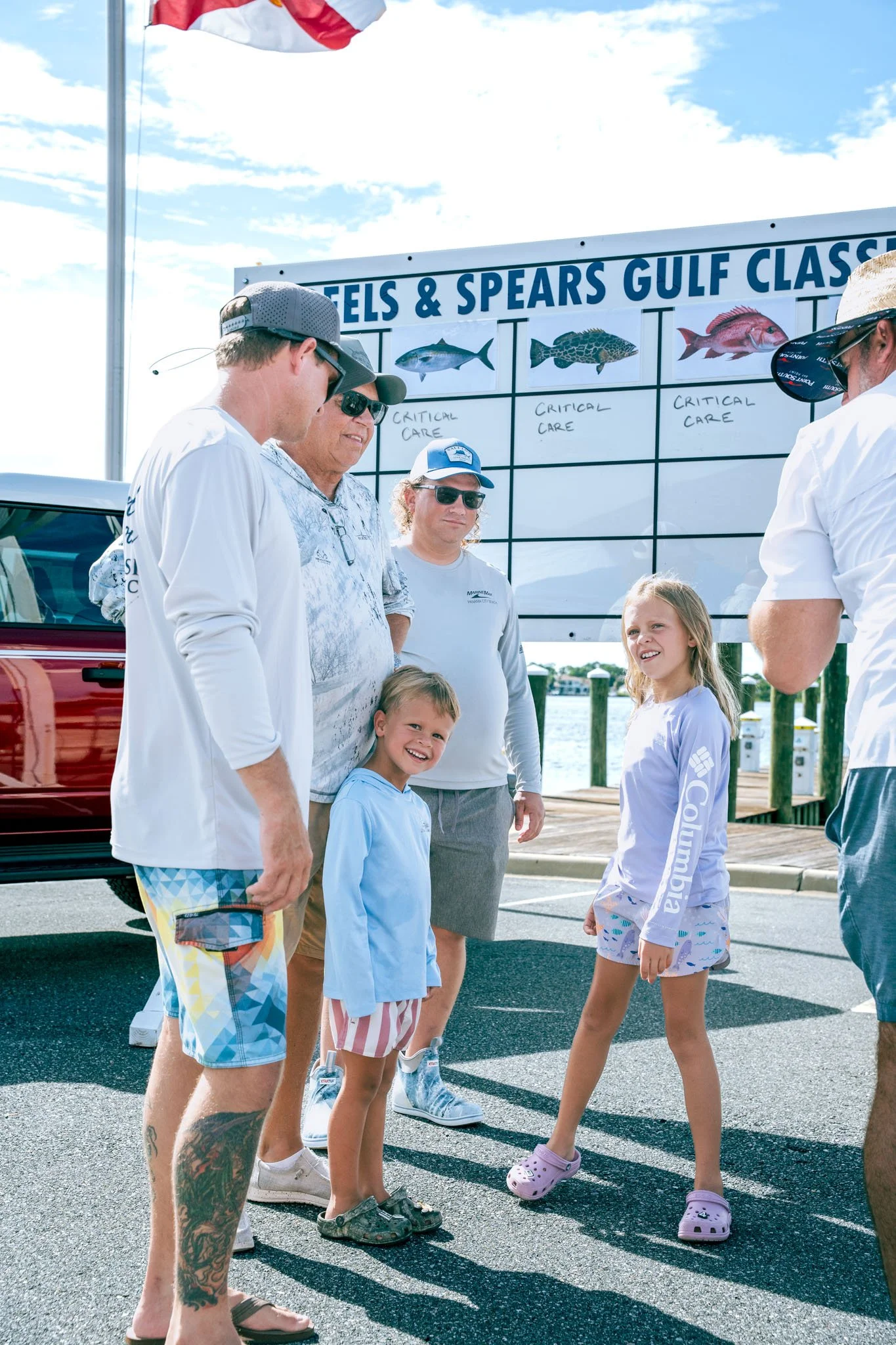 A group of people, including children and adults, standing near a sign that shows images of different fish and the text "TIELS & SPEARS GULF CLASS". The background features a body of water, a dock, and an American flag flying in the sky.