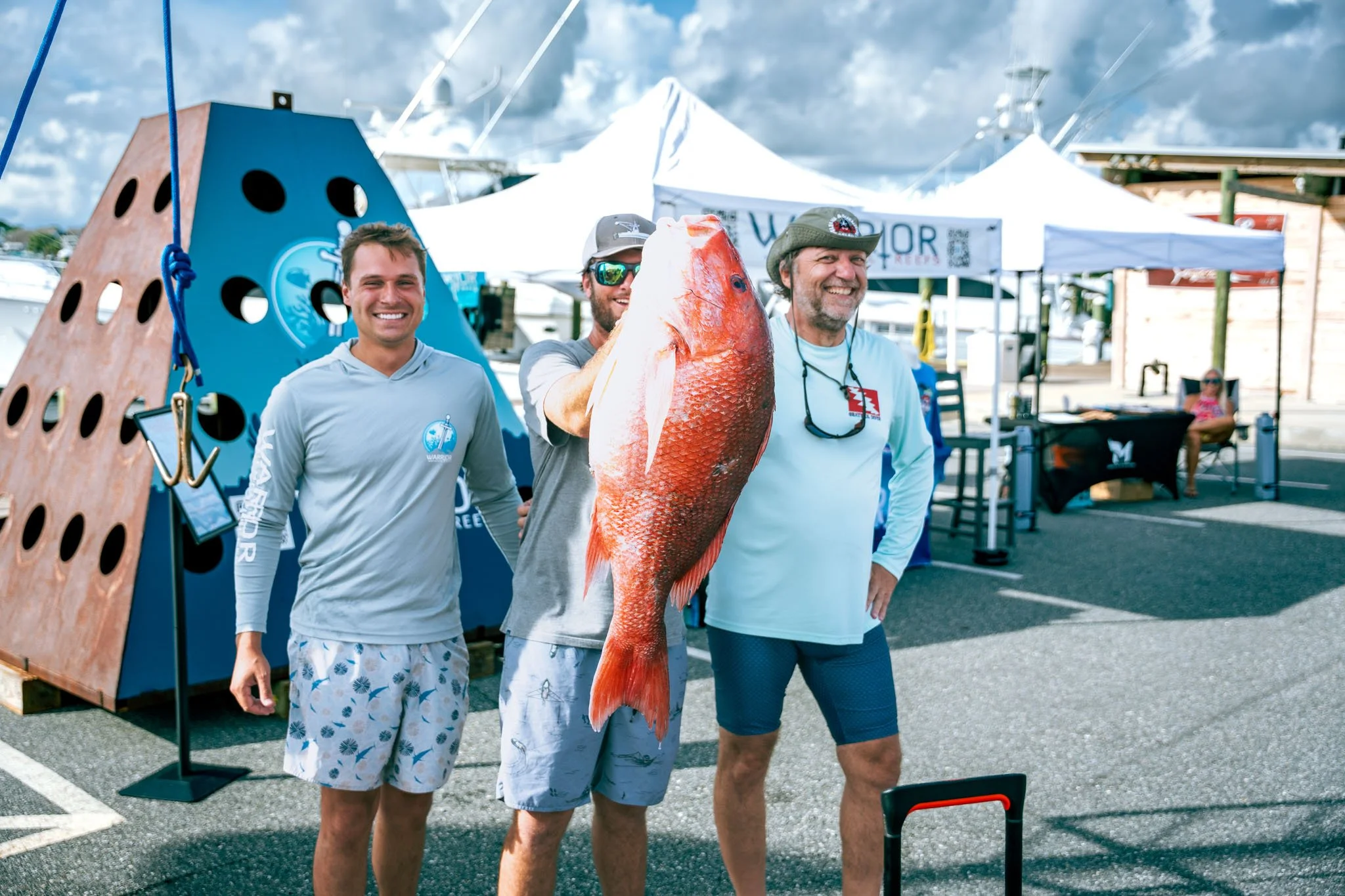Three men standing outdoors at a fishing event at Point South Marina - Bay Point (Panama City Beach), holding a large red fish, with tents and other event structures in the background, under cloudy skies.