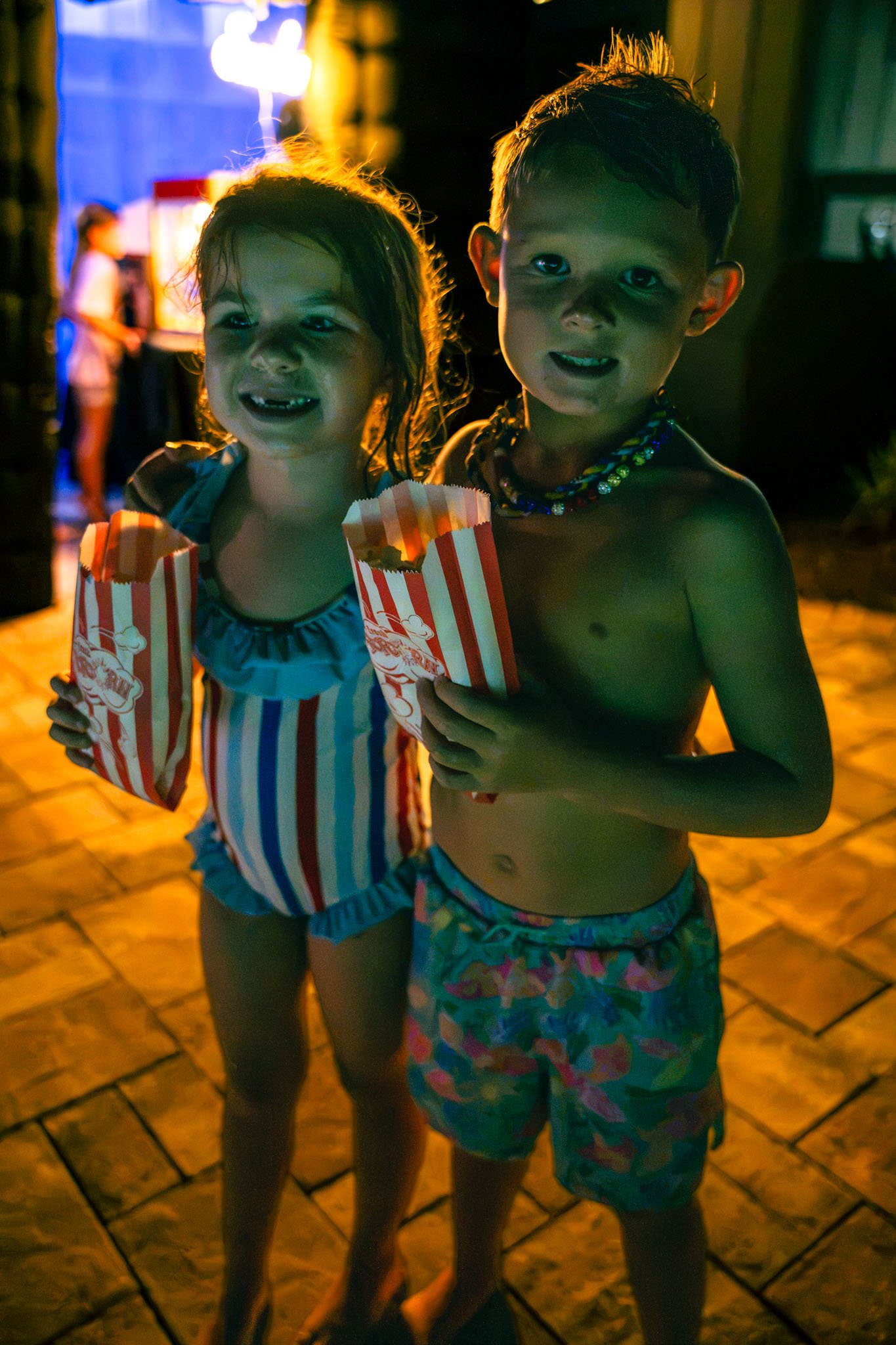 Two young children, a girl and a boy, standing together at night, holding striped popcorn bags, with a person in the background near a brightly lit area.