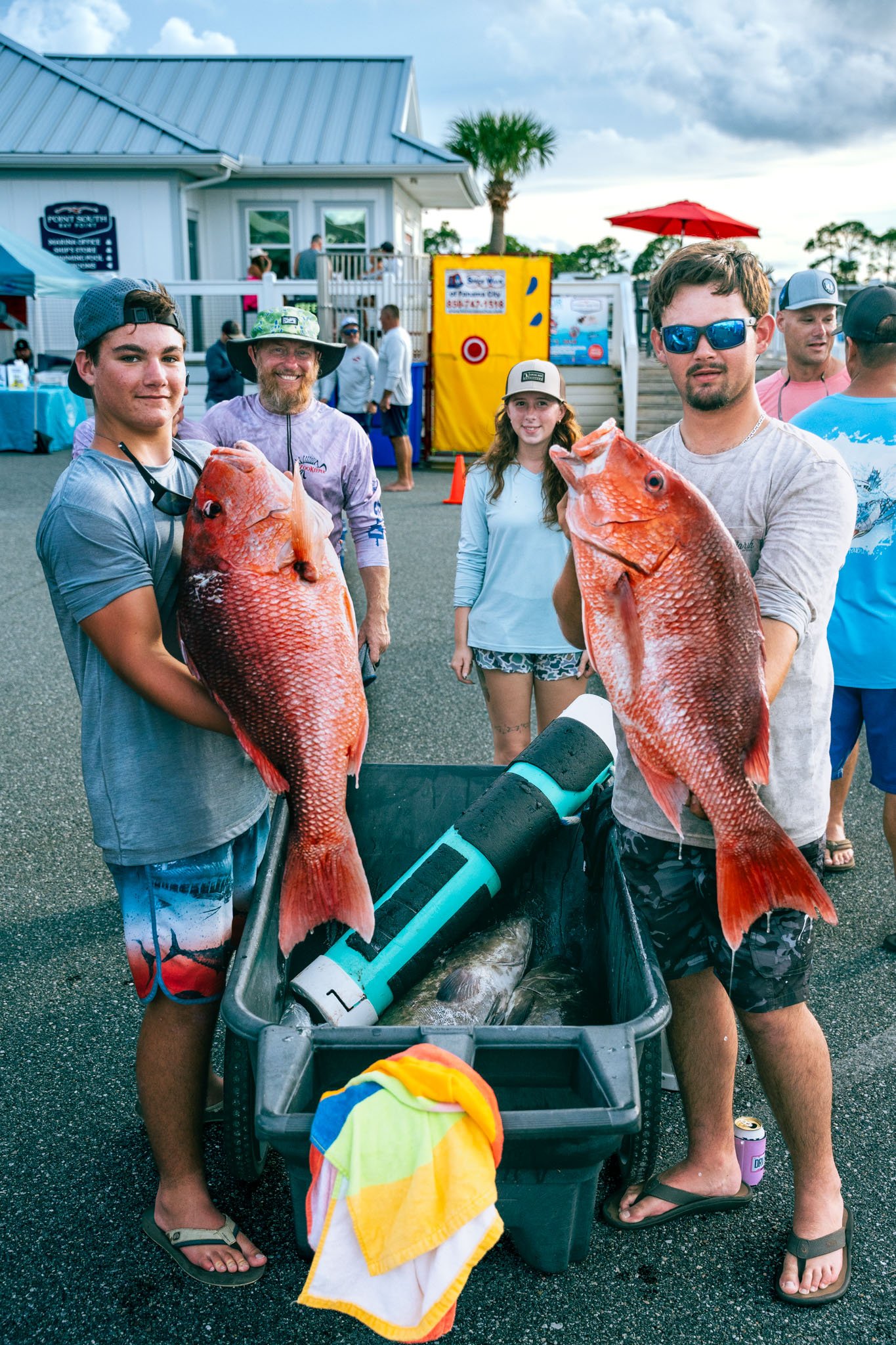 A group of five people holding large red snapper fish at an outdoor event, with a cart filled with more fish and a water cannon in front. In the background, there are tents, a building, and other people, under a cloudy sky.