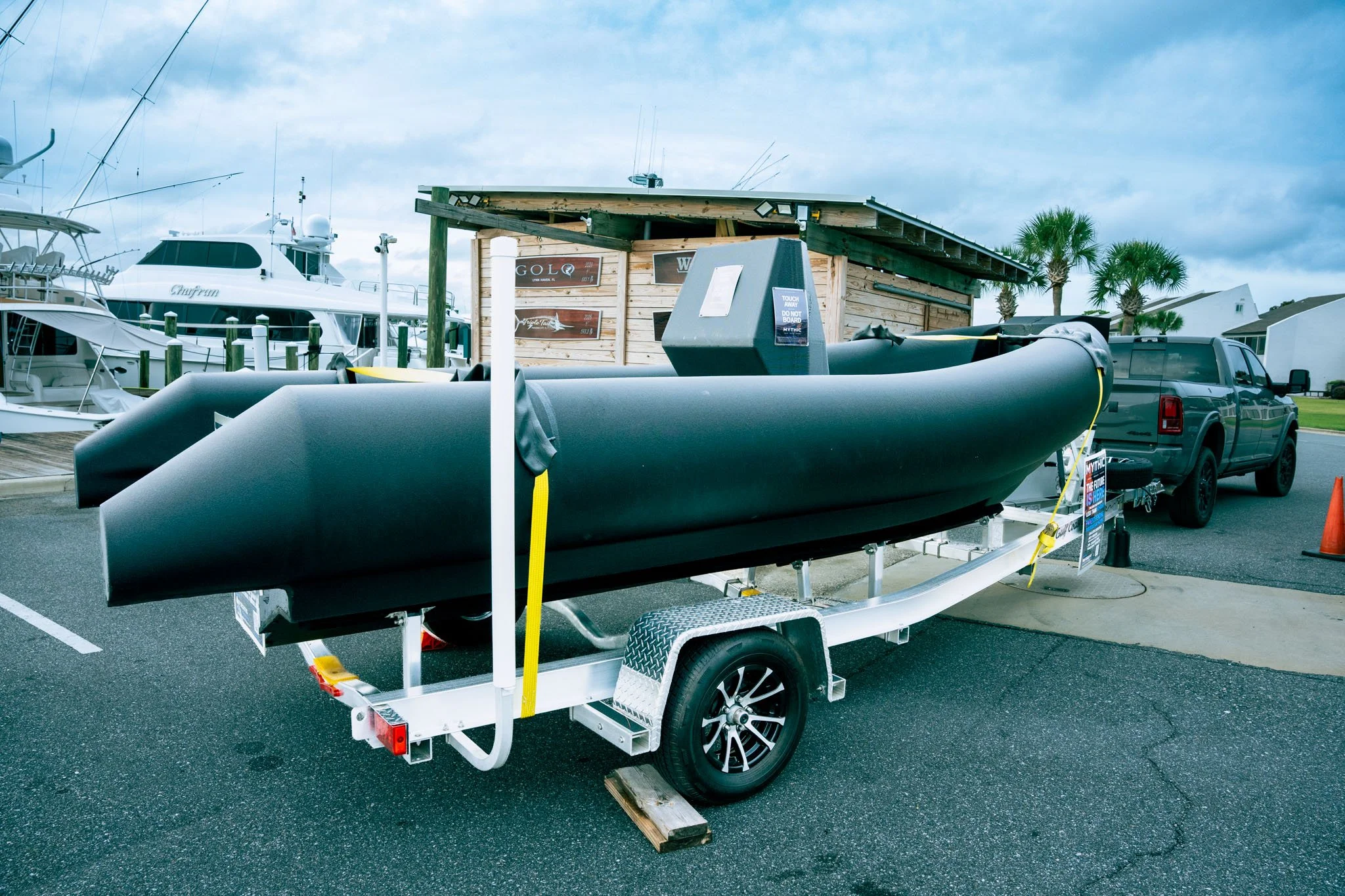 A boat trailer with a black inflatable boat, boats docked at a marina, and a small wooden building with restaurants signs in the background, along with palm trees and a cloudy sky.