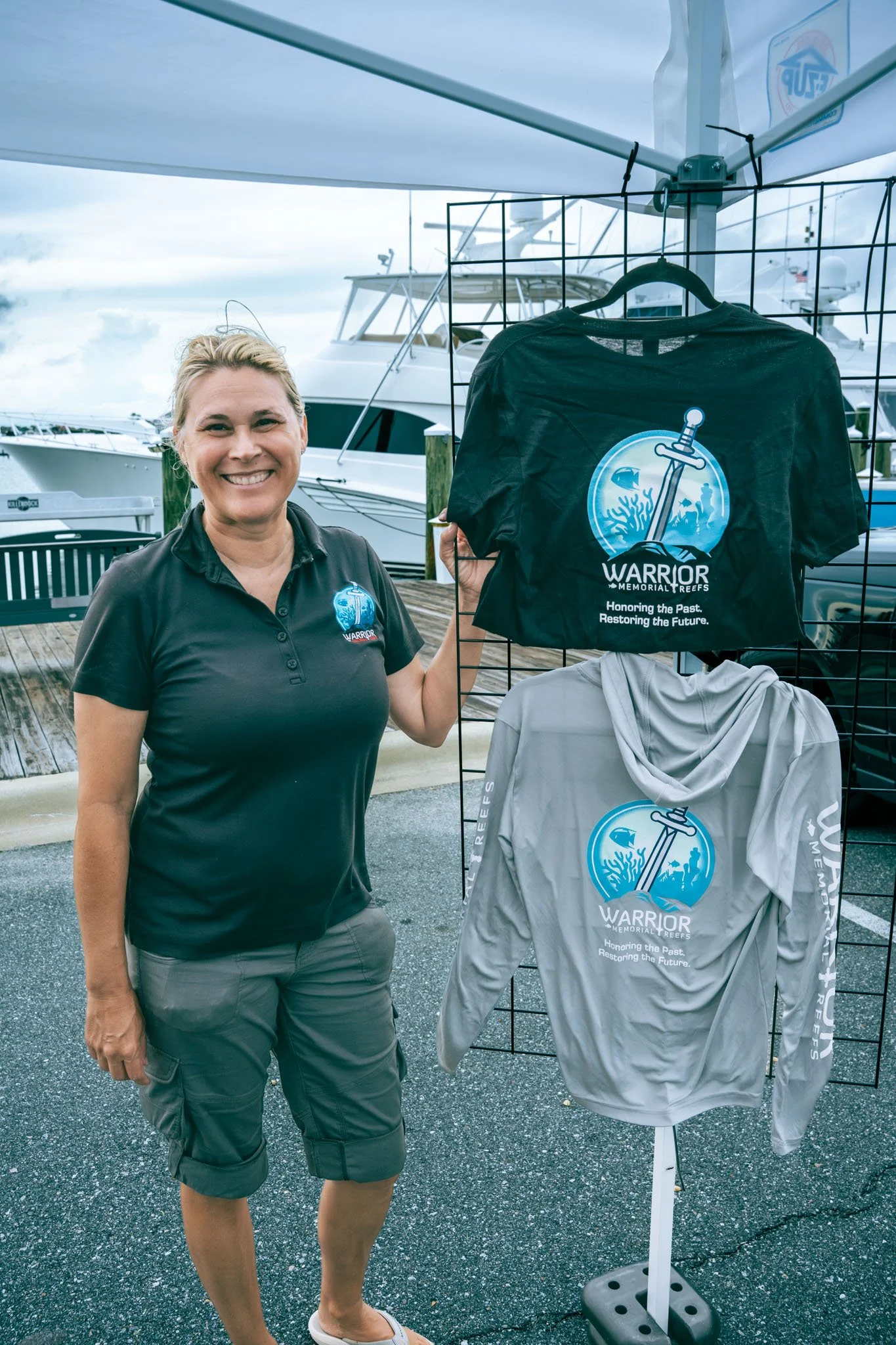 Woman smiling at a marina next to a display of t-shirts with a logo that reads 'Warrior Memorial Reefs' and the slogan 'Honoring the Past. Restoring the Future.' She is wearing a black polo shirt with the same logo, gray shorts, and white slip-on sho