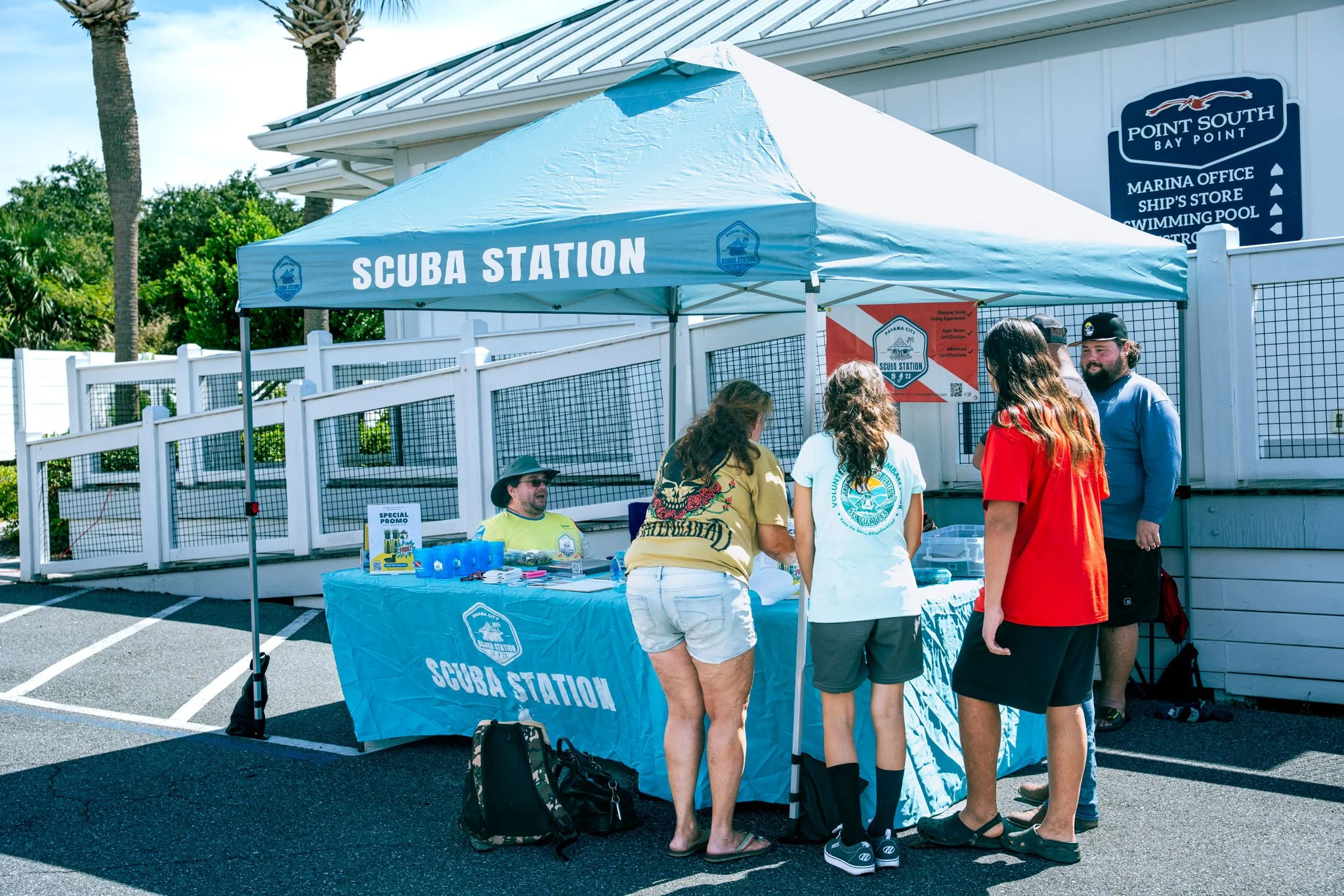 People gather around a blue tent with a sign that reads 'Scuba Station' at an outdoor event near a white building with a sign pointing to various locations such as the marina office, ship's store, swimming pool, and restroom. The area is sunny with p