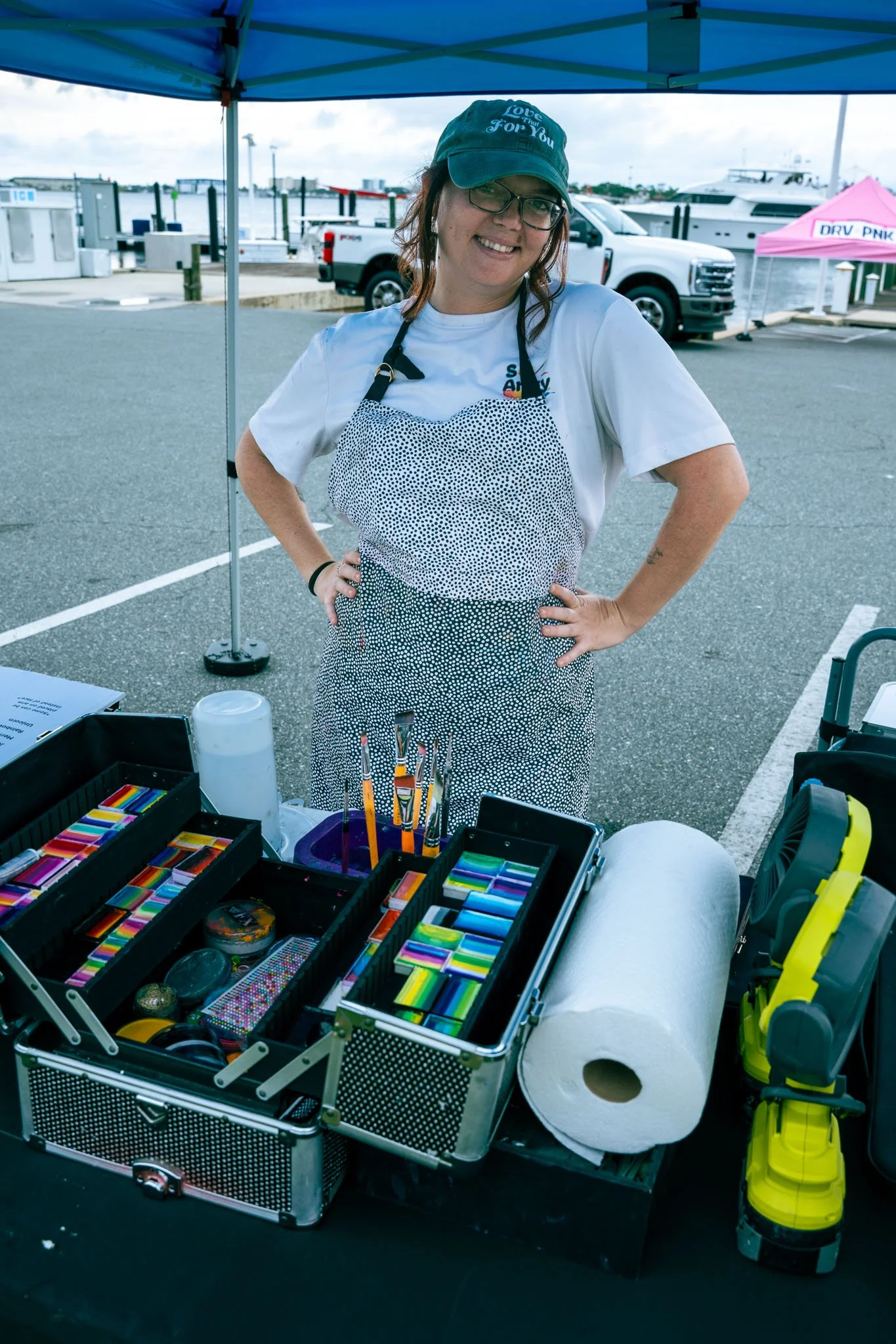 Smiling woman wearing glasses, a baseball cap that says 'Love That For You,' and an apron, standing behind a table of various face paint art supplies, including colorful pastels, brushes, and tape, outdoors near boats and trucks.