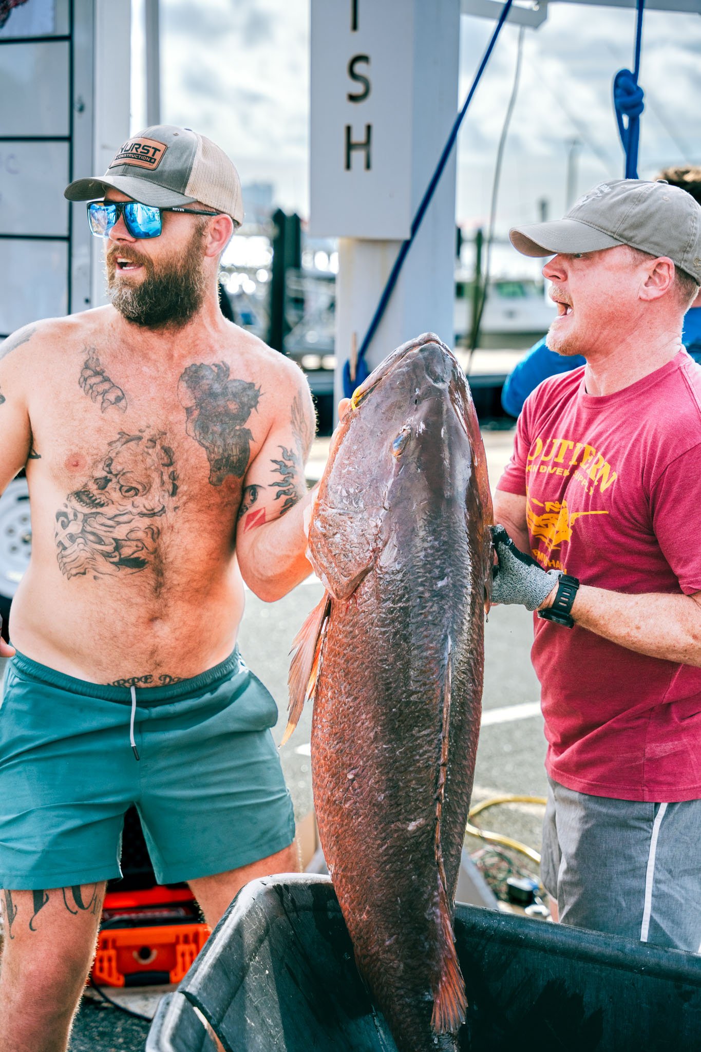 Two men are holding a large fish at a dock, one shirtless with tattoos, sunglasses, and a cap, and the other in a red t-shirt and cap, with boats and a cloudy sky in the background.