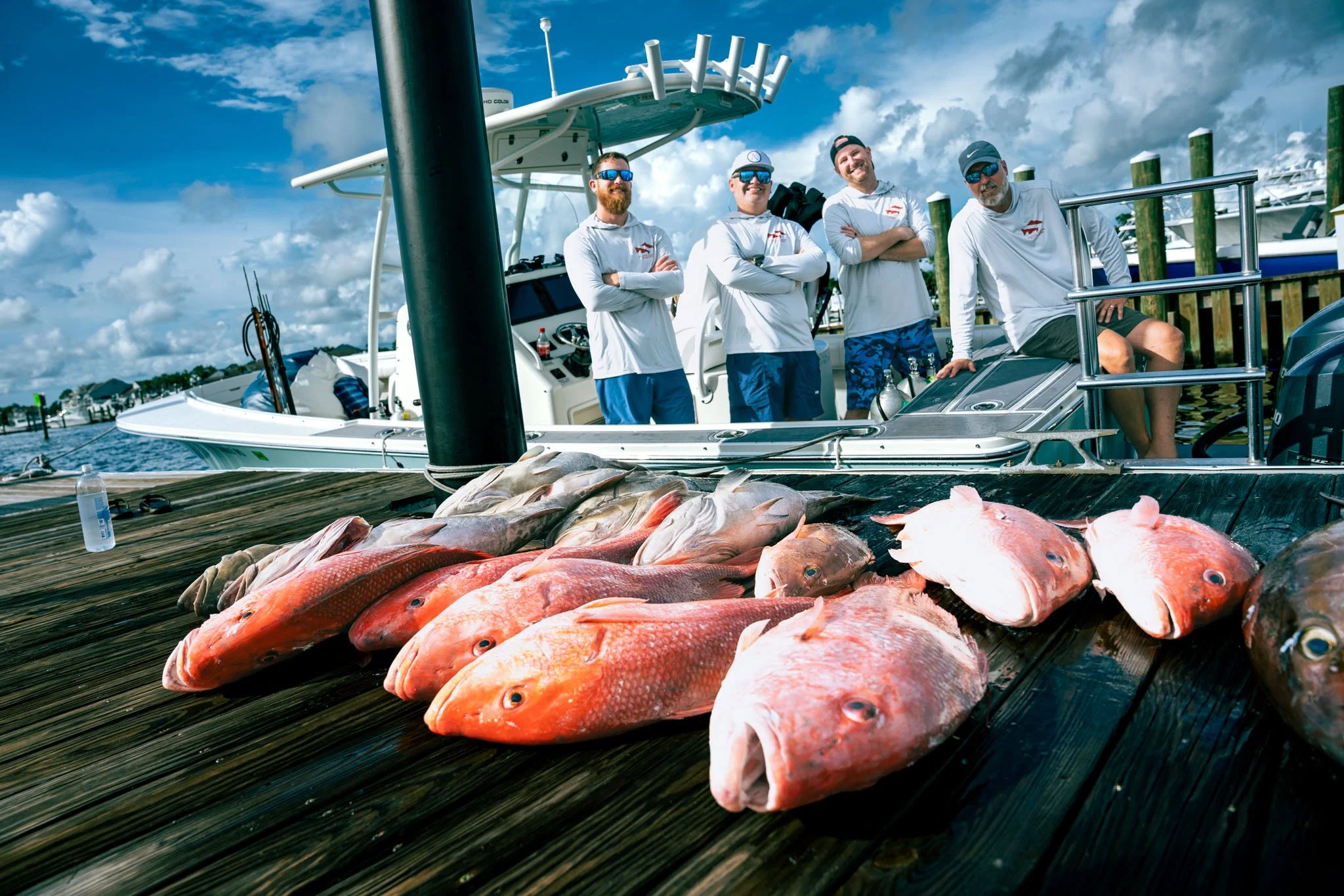 A group of four men standing on a dock with a boat in the background, posing with arms crossed, next to a display of various freshly caught fish laid out on a wooden surface. The sky is partly cloudy with a marina in the distance.