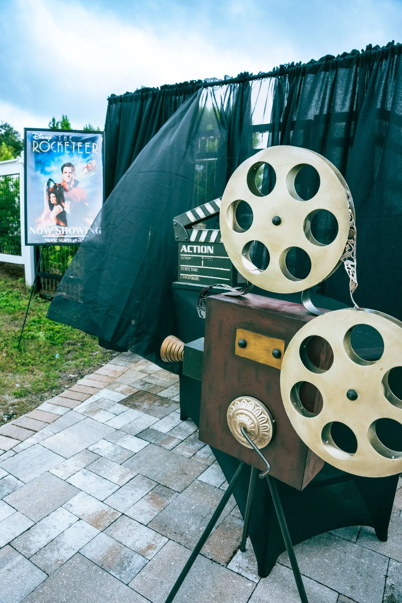 Film reels and a movie clapperboard on display outdoors during daytime, with a movie poster in the background for Disney's 'The Rocketeers'.