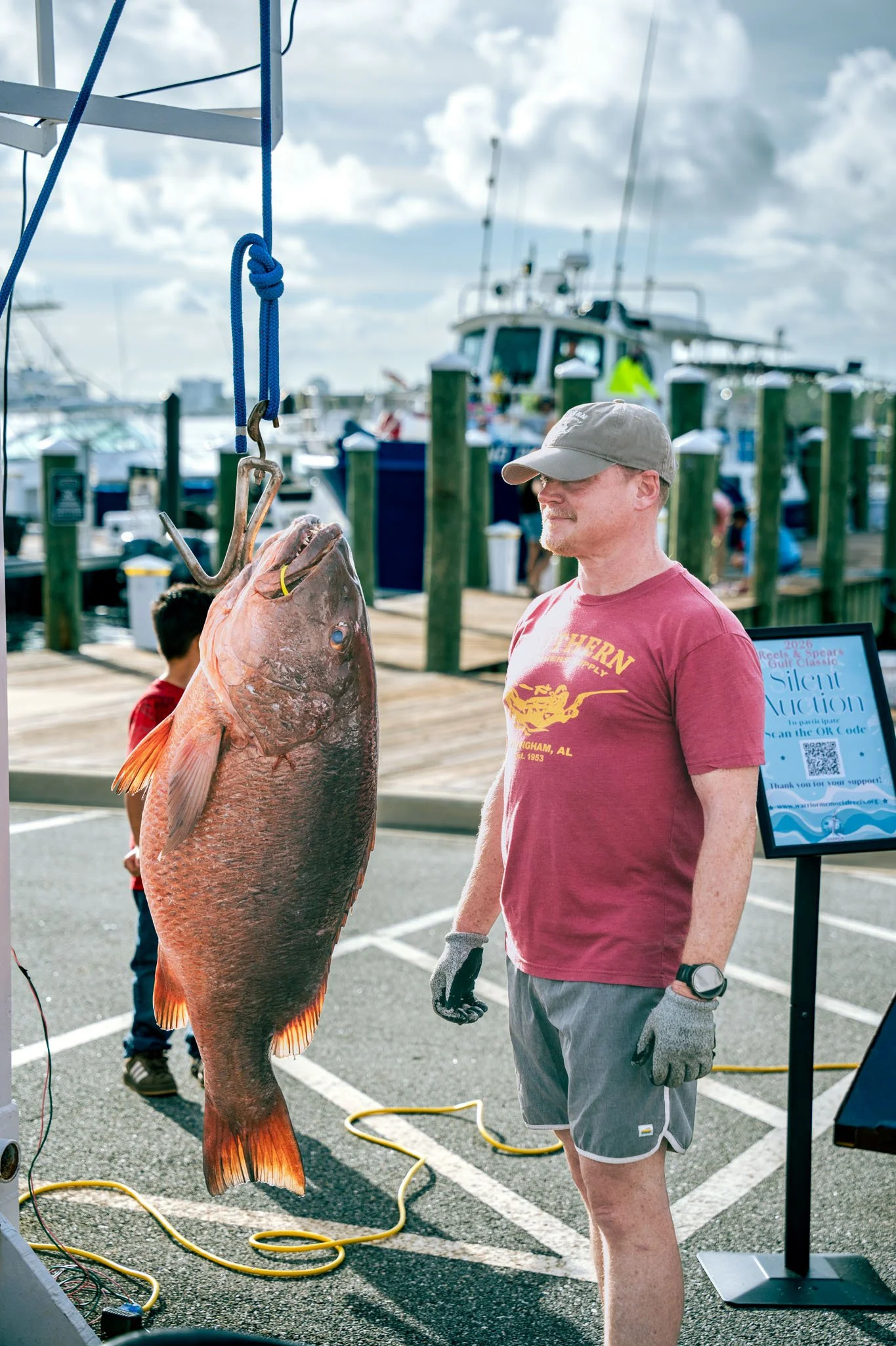 A man wearing a beige cap, gray shorts, and gloves stands next to a large cubera snapper hanging from a scale. Other boats are visible in the background at a marina.