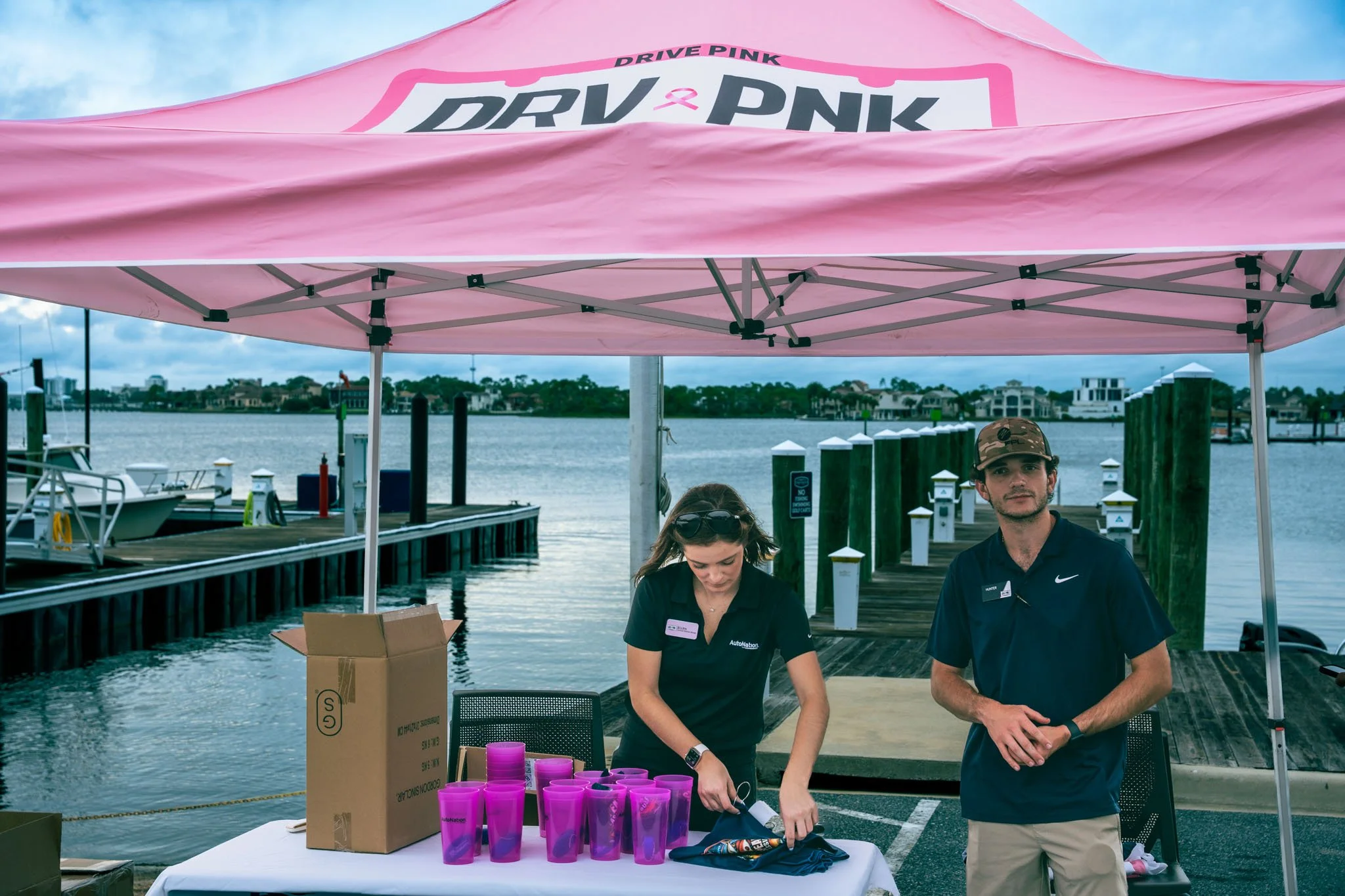 Two event staff members standing under a pink canopy at a waterfront location, with one woman organizing purple cups and a man looking towards the camera, nearby tables, boxes, and a marina in the background.