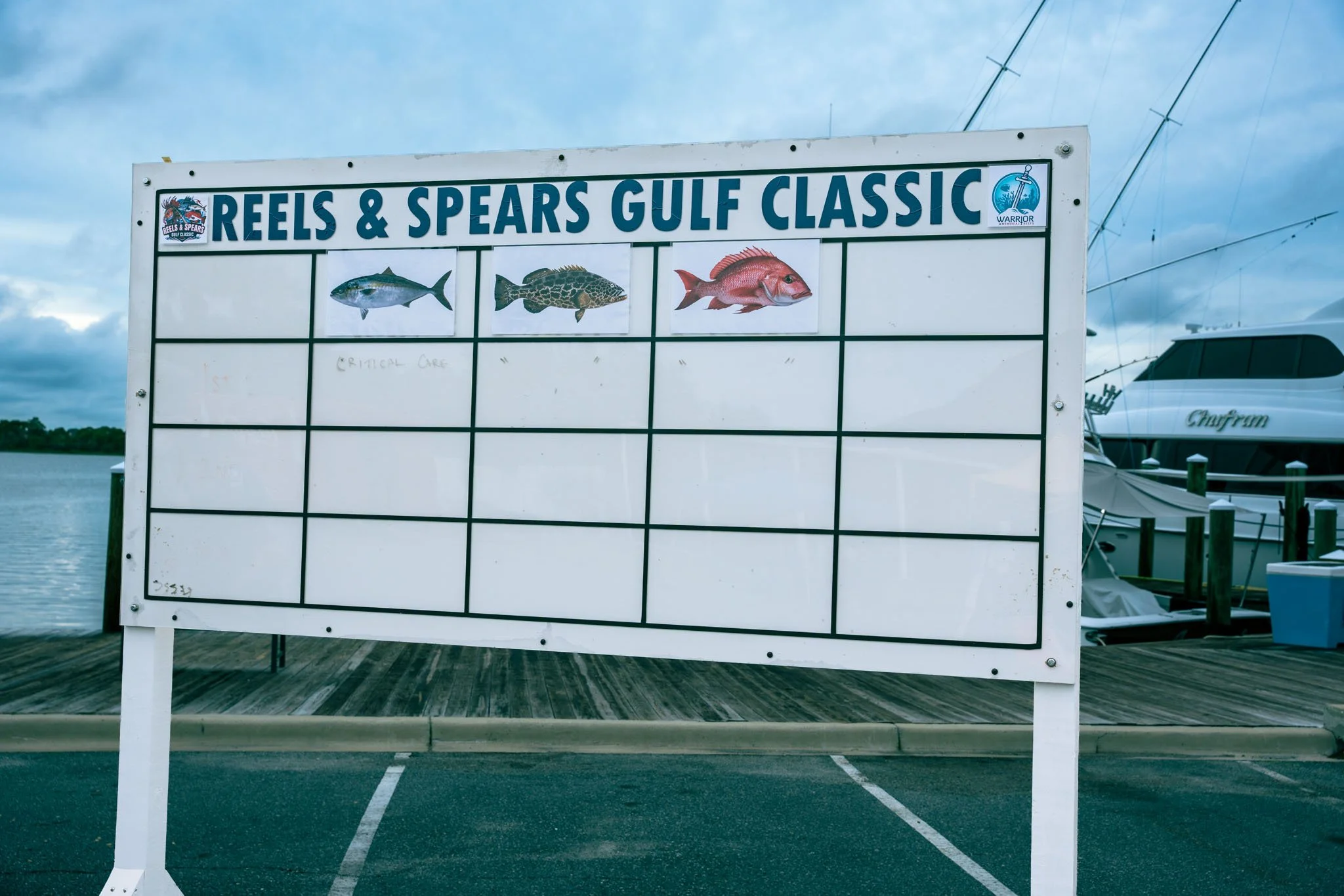 Sign with pictures of three fish and text reading 'Reels & Spears Gulf Classic' at a marina with boats, water, and cloudy sky in the background.