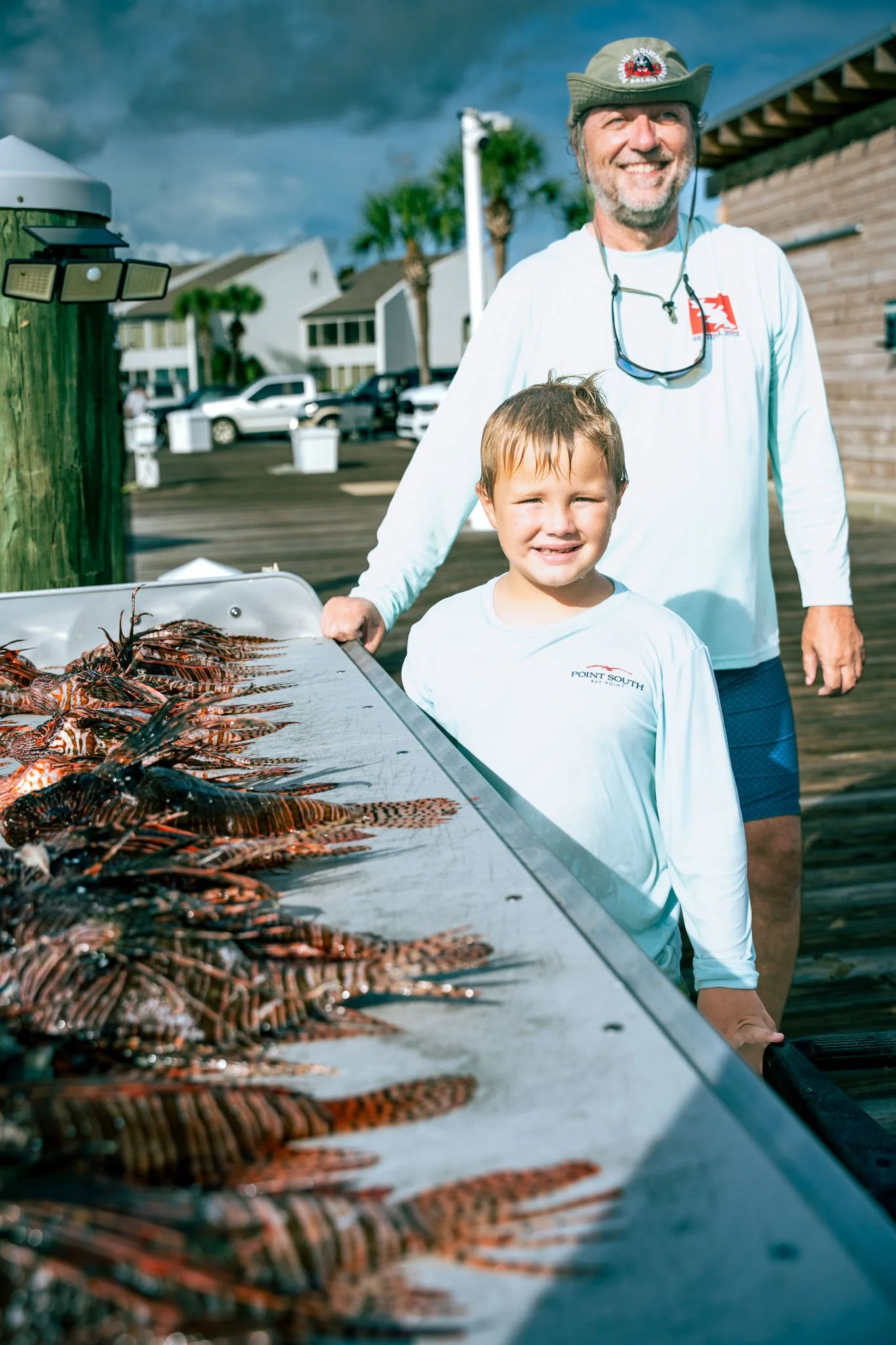 A man and a child standing behind a grill filled with cooked lobster on a dock or pier, with parked cars, houses, palm trees, and a cloudy sky in the background.