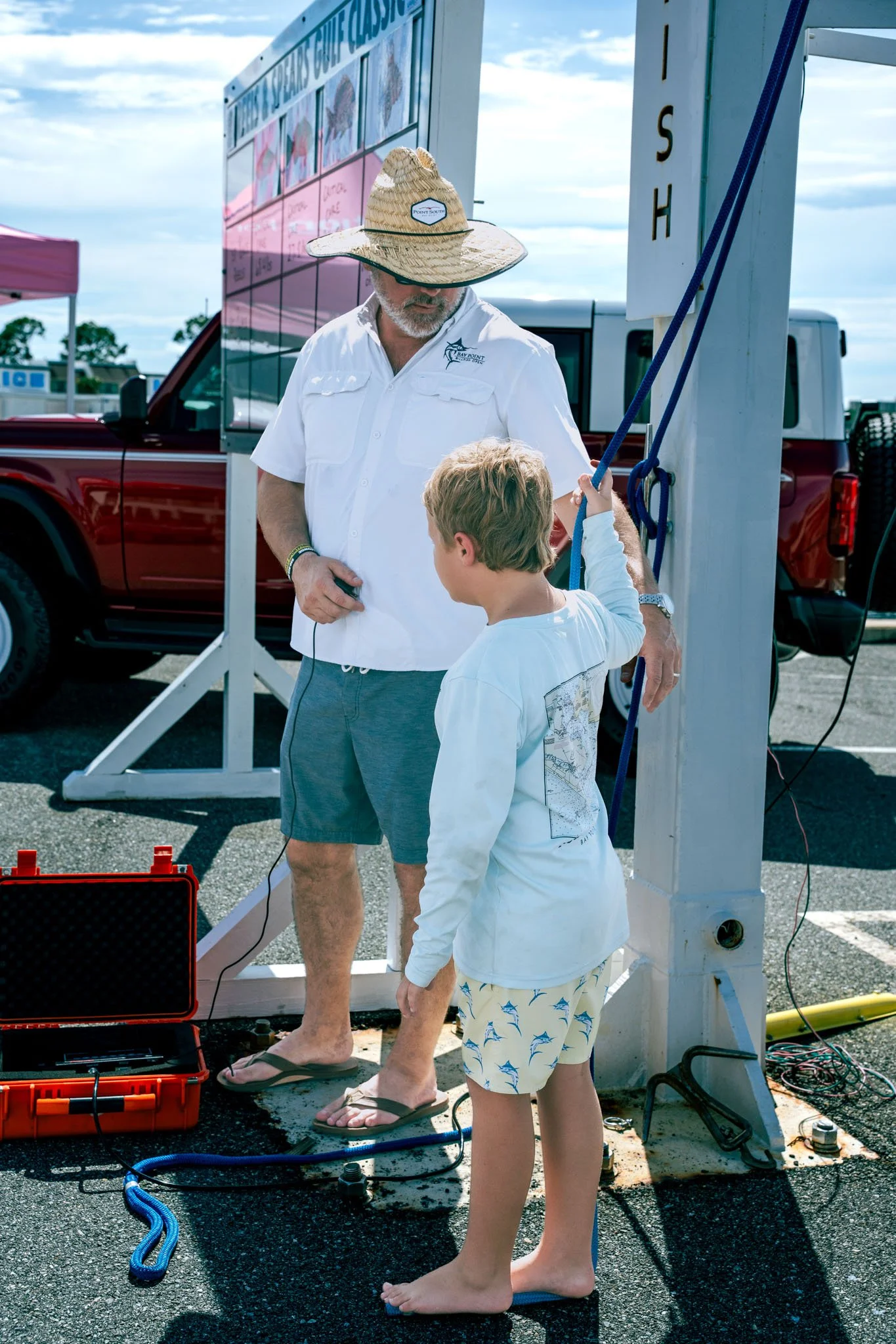 A man and a young boy with blond hair at a fishing check-in booth by the water, with a red truck in the background. The man is wearing a straw hat, white shirt, and shorts, and the boy is in a white long-sleeve shirt and swim trunks with fish print. 
