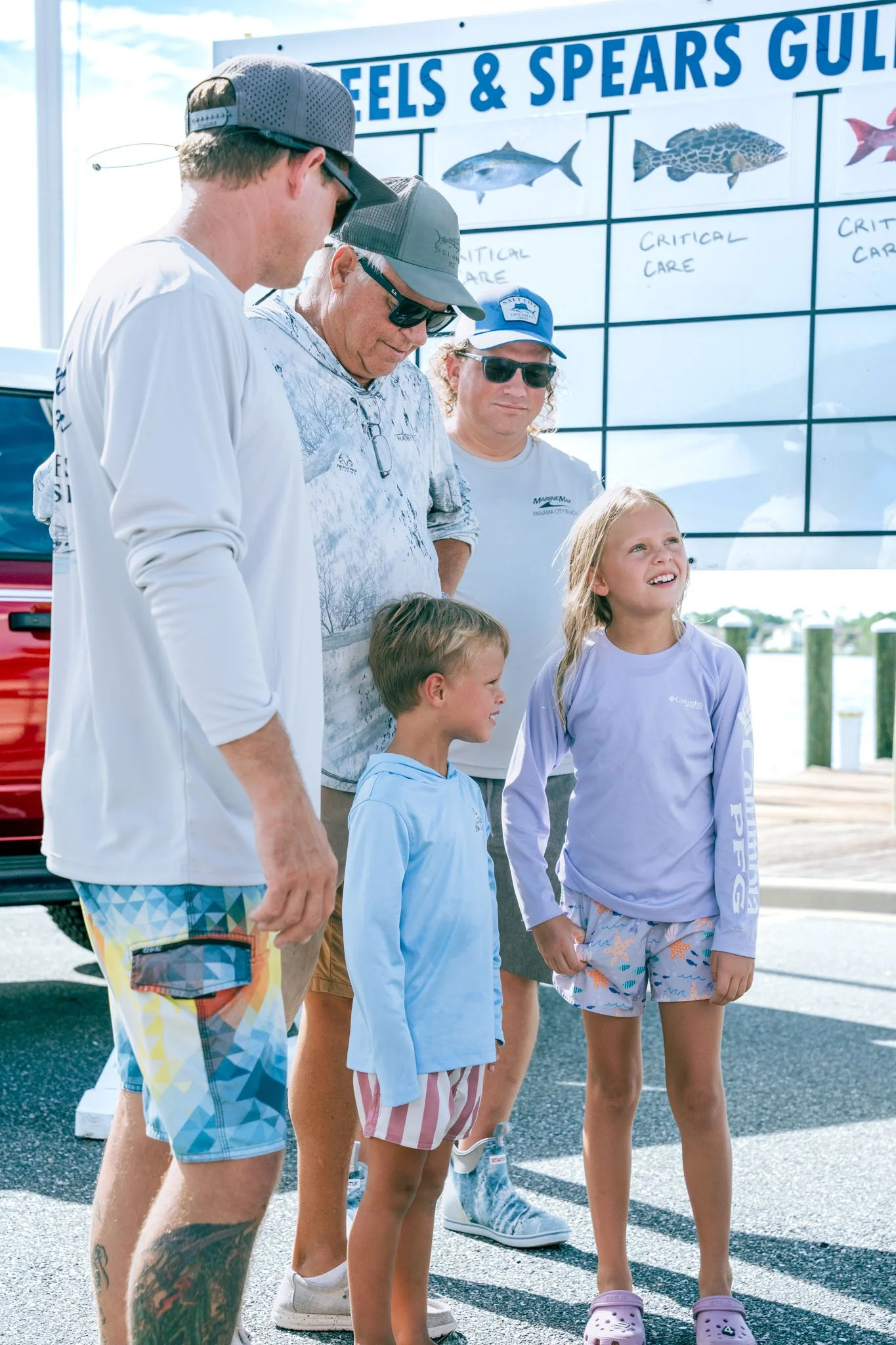 Group of people, including children and adults, standing in a line outdoors near a board with fish illustrations and descriptions at a marina or fishing area.