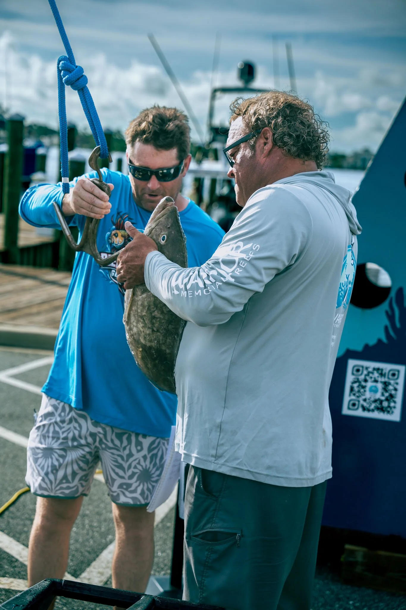 Two men are holding a fish together on a dock at Point South Marina - Bay Point (Panama City Beach), with one man using a tool to hold the fish's mouth open and the other supporting it. They are near boats and dock posts, with a cloudy sky overhead.