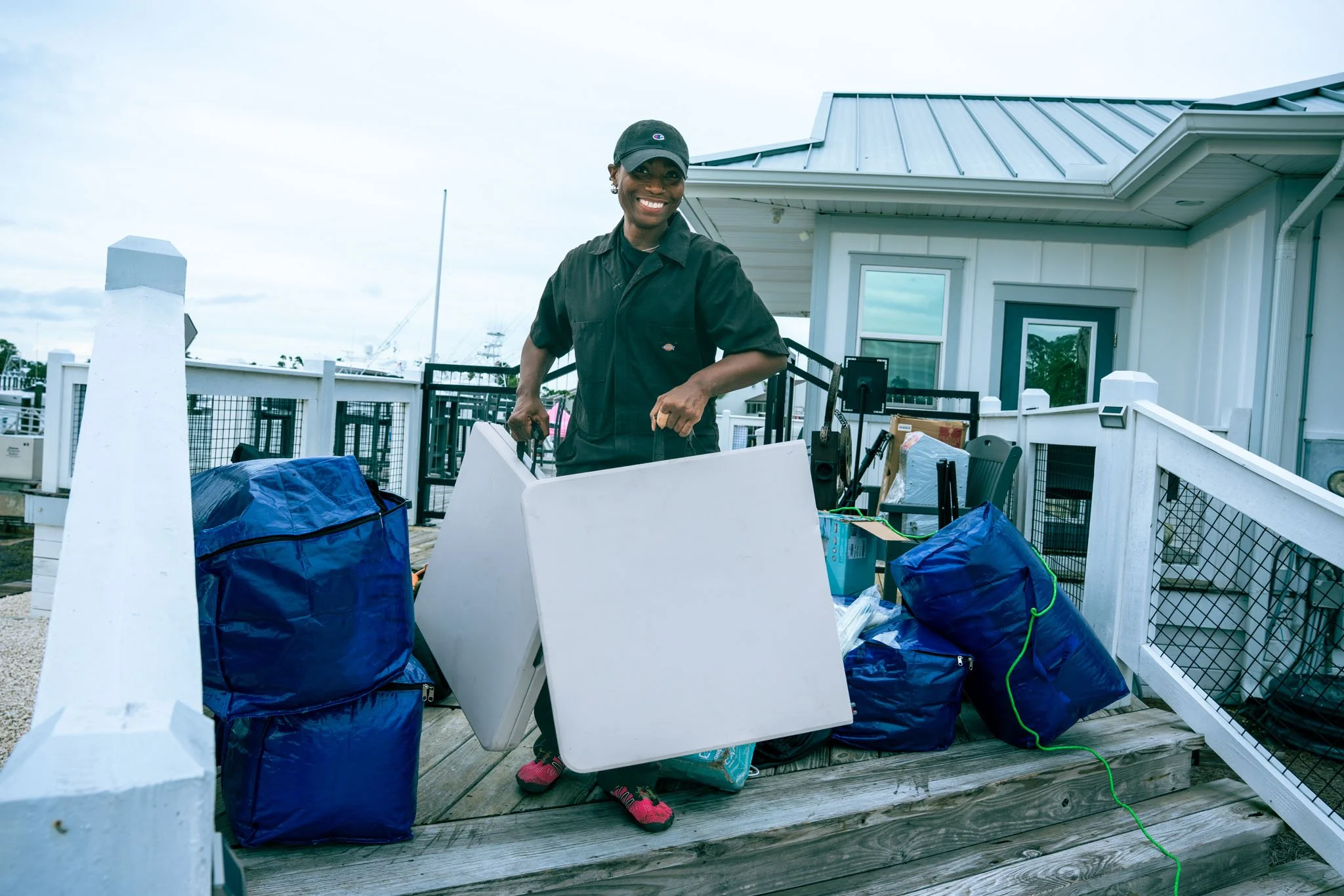 A smiling woman at Point South Marina - Bay Point (Panama City Beach) wearing a black baseball cap and black shirt stands on a porch surrounded by blue bags and white furniture, with a house and cloudy sky in the background.