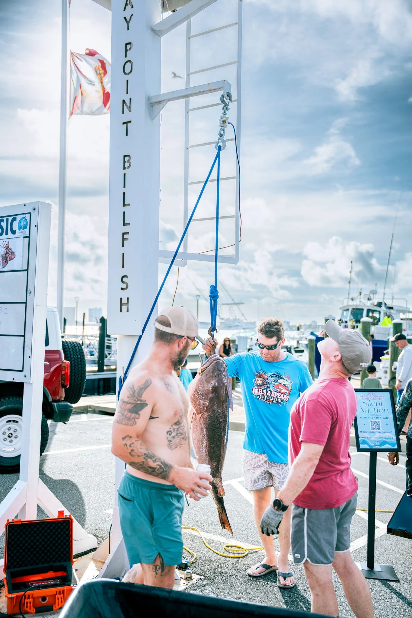People at the Warrior Memorial Reefs Reels and Spears Fishing Tournament event at Point South Marina - Bay Point (Panama City Beach), some examining a large cubera snapper being held by a man, with boats and a cloudy sky in the background.