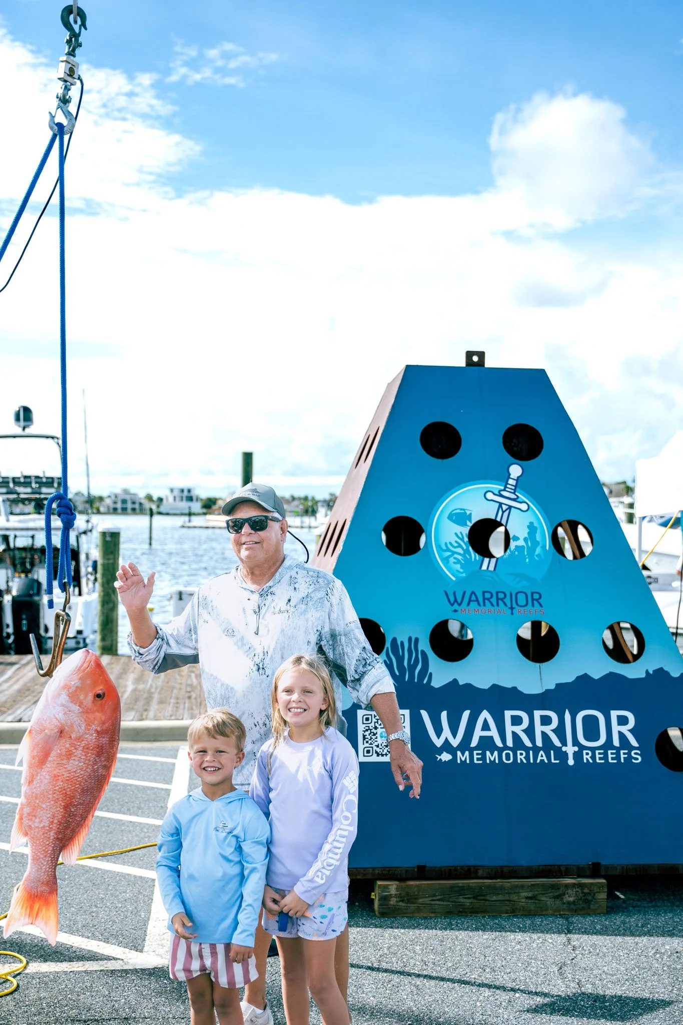 A group of people at Point South Marina - Bay Point (Panama City Beach) with a large whale tail sculpture and a sign that reads 'Warrior Memorial Reefs'. Children and an adult smiling, with a large fish hanging nearby.