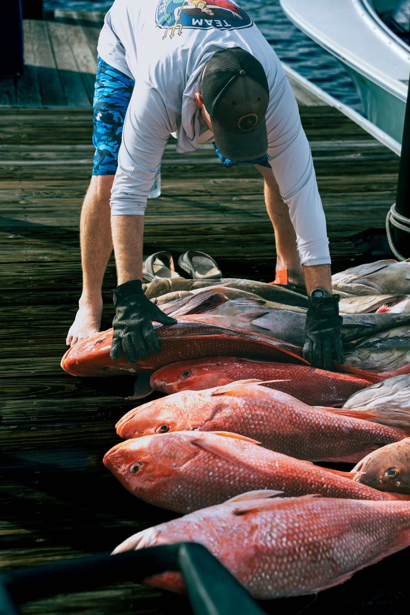 A man bending over a large catch of red fish, preparing them on a wooden dock during daytime. He is wearing a black cap, white long-sleeve shirt, and blue camouflage shorts. The background shows water and a boat.