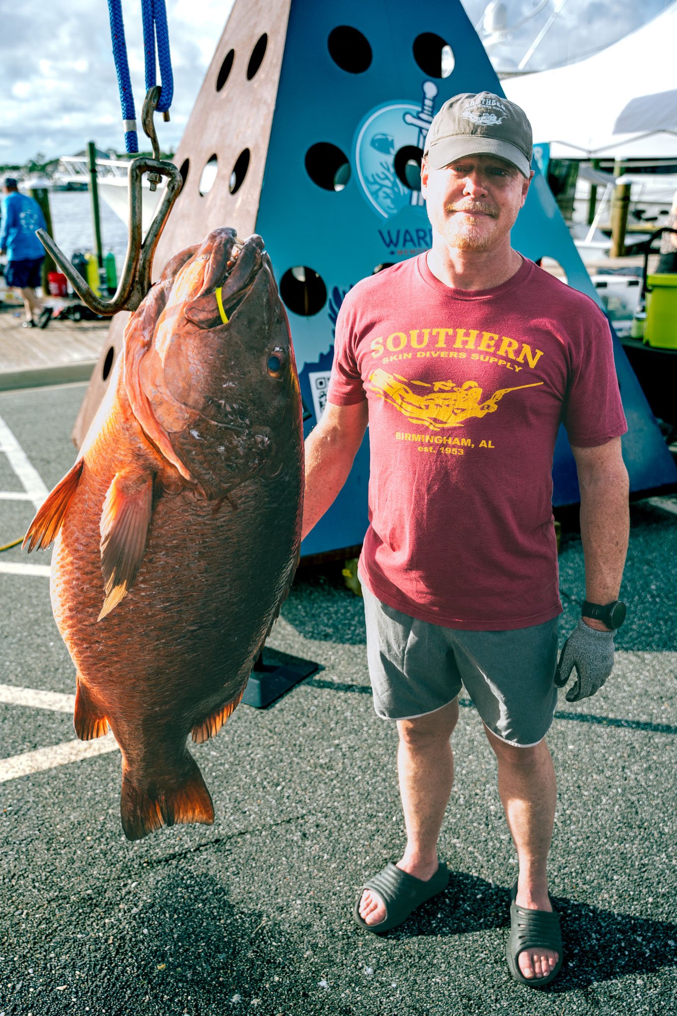 A man in a red t-shirt, gray shorts, flip-flops, and a beige cap is standing on a paved area  A large cubera snapper is next to him, hanging from a metal hook and a blue rope. In the background, there are boats, tents, and other people.