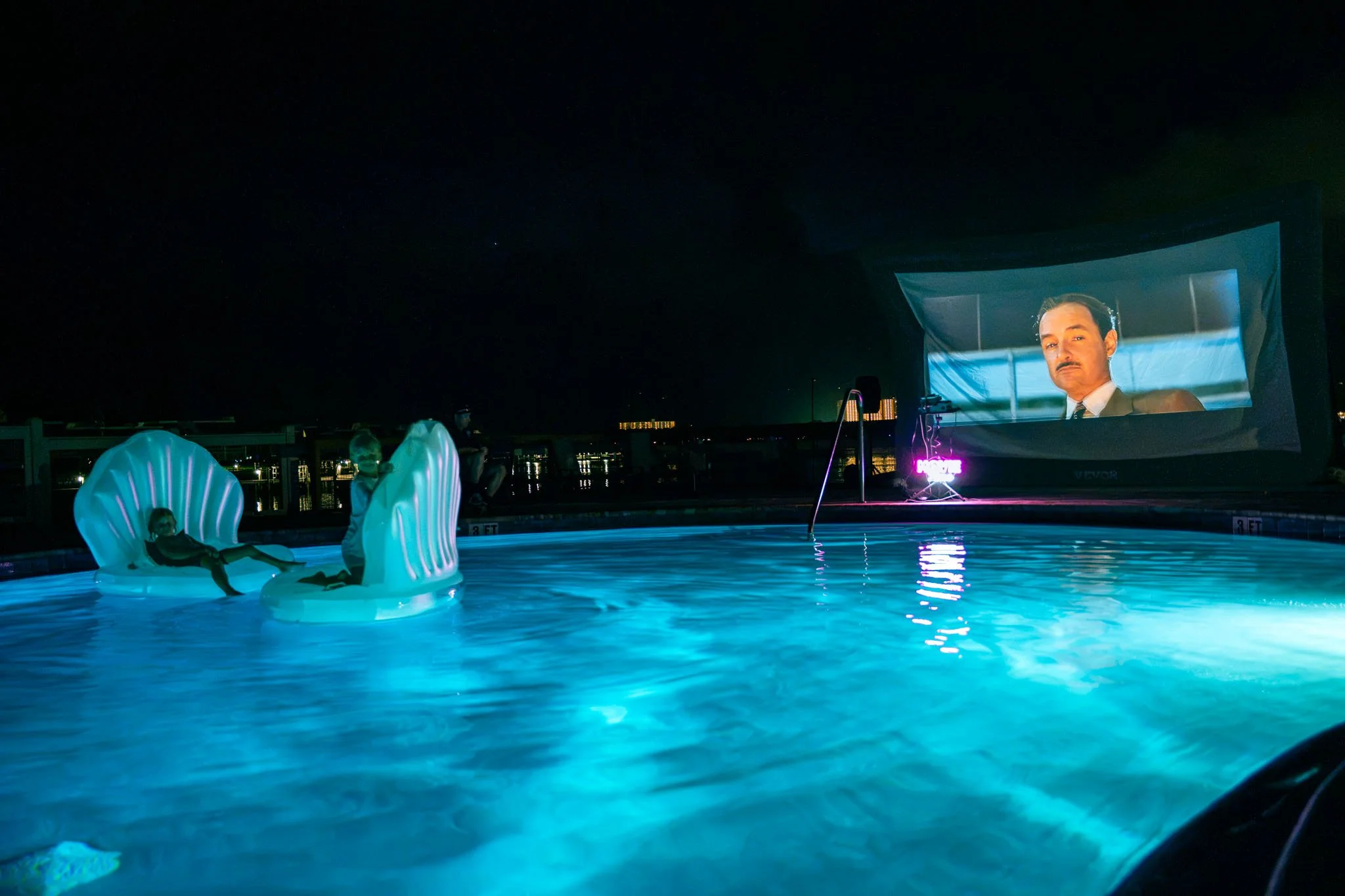 Nighttime scene at Point South Marina - Bay Point pool showing a large cinema screen displaying a man with a mustache in a suit. The pool is illuminated with blue lighting, and there are inflatables and people relaxing on the poolside chairs.
