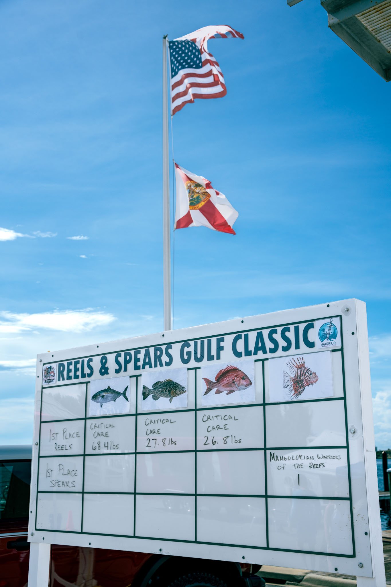A whiteboard displays the results of a fishing competition at the Reels & Spearsgulf Classic, with photos of different fish and their critical care weights. Two flags, the United States and Florida state flags, are flying above the whiteboard against
