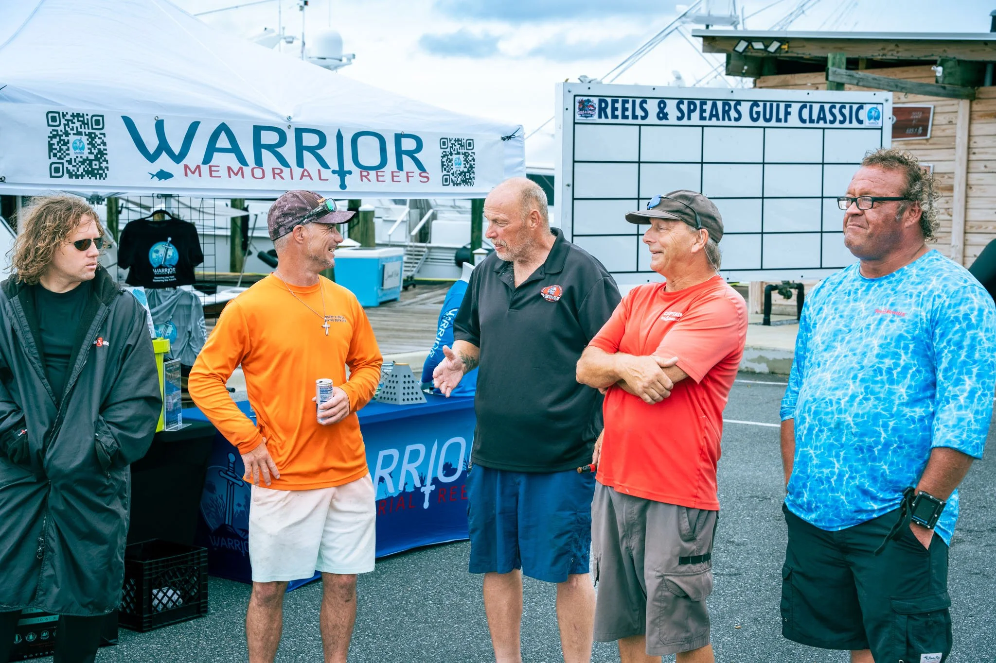 Group of men standing together outdoors near a booth with signs for Warrior Memorial Reefs and Reels & Spears Gulf Classic. The men are engaged in conversation, some smiling and some serious, wearing casual clothing including hats and sunglasses.