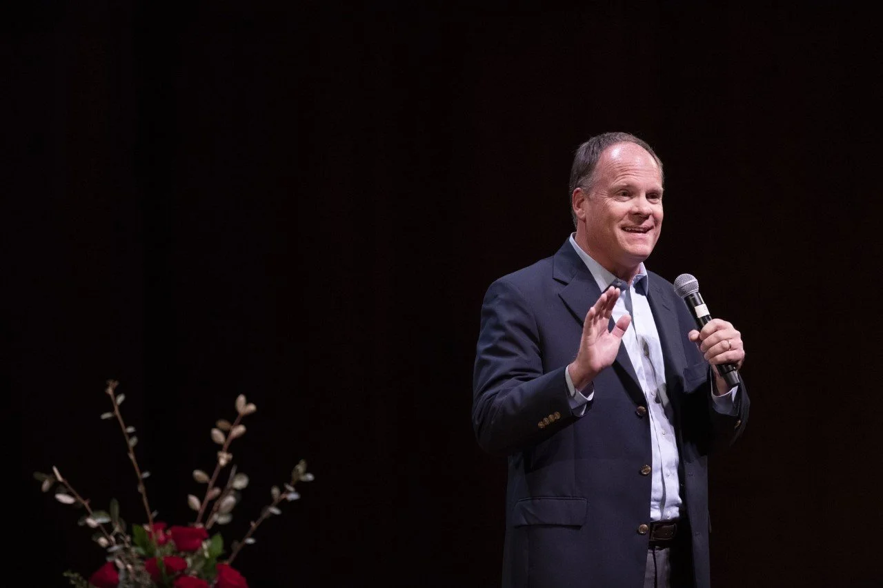 A man in a dark suit holding a microphone, speaking on stage with a dark background and a floral arrangement to his left.
