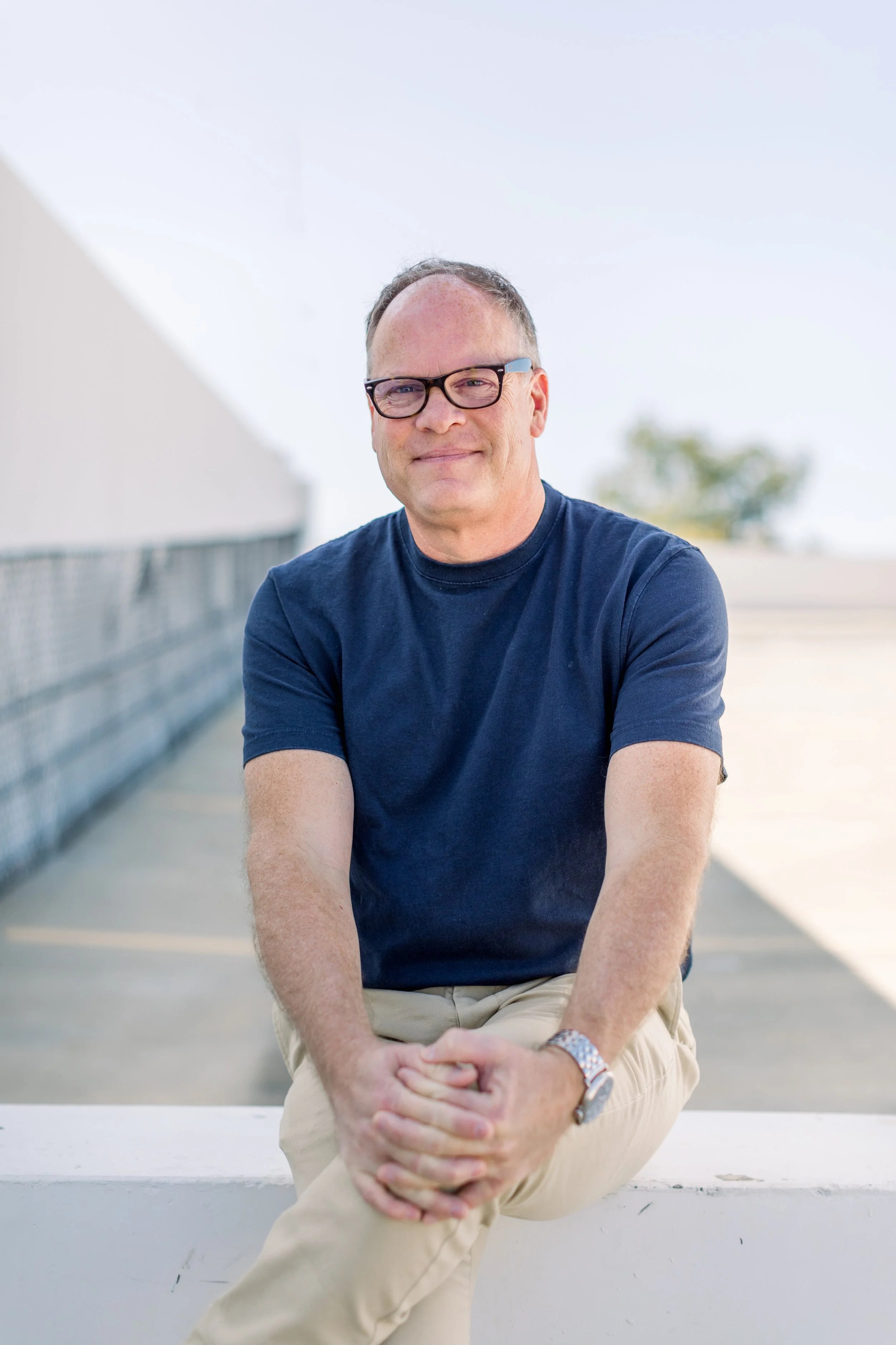 Man in glasses wearing navy T-shirt and khakis sitting outdoors on a white ledge, with a blurred background of trees and a building.