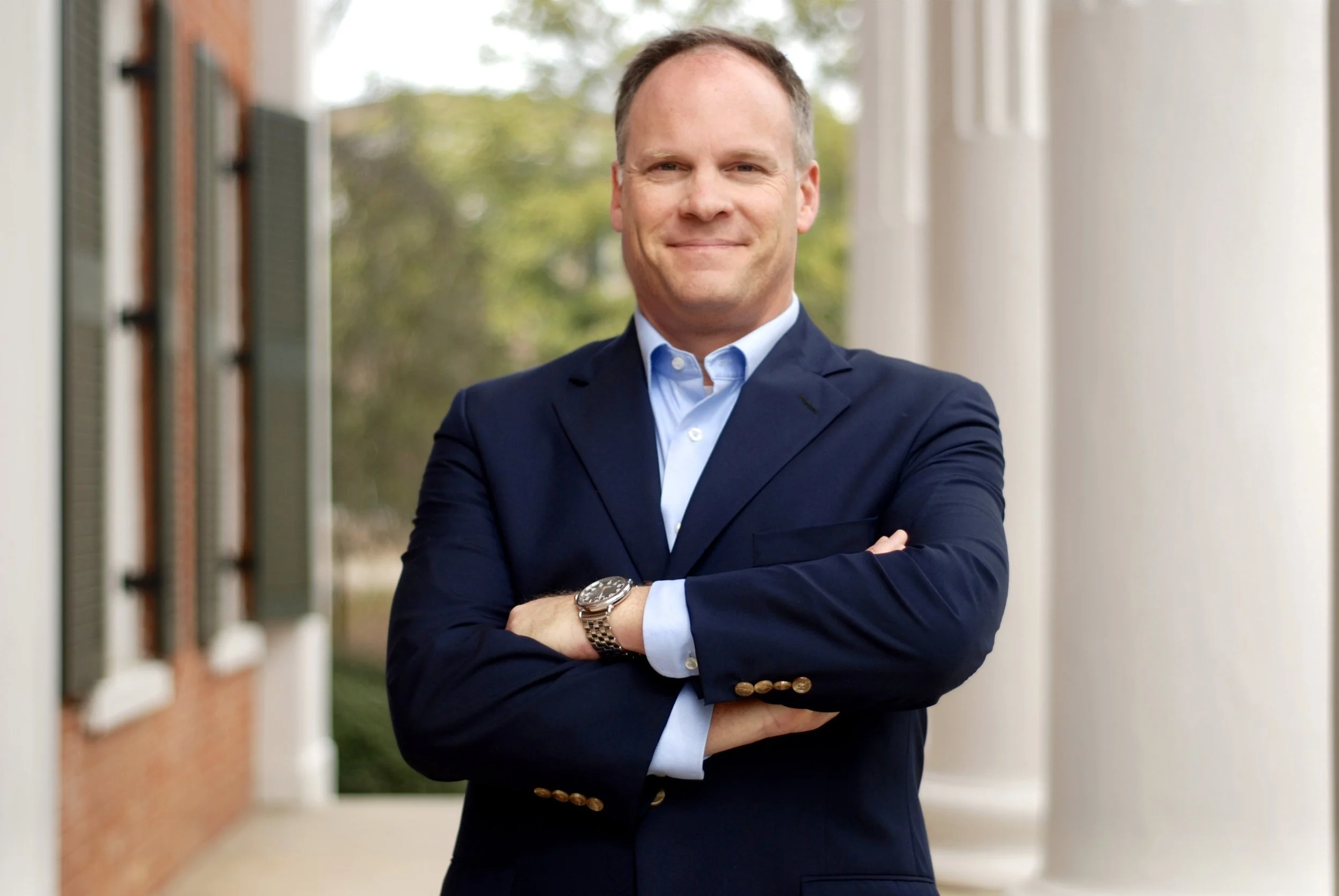 A man wearing a navy blue suit, light blue shirt, and watch, standing outdoors with arms crossed, smiling at the camera.