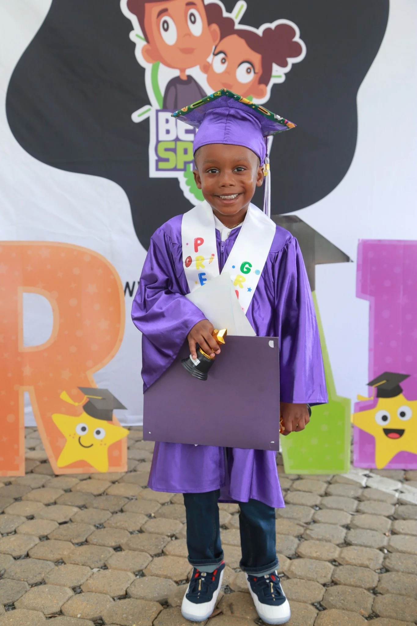 Young child dressed in a purple graduation cap and gown, holding a diploma, standing on a cobblestone surface in front of a cartoon-themed backdrop with colorful letters and stars.