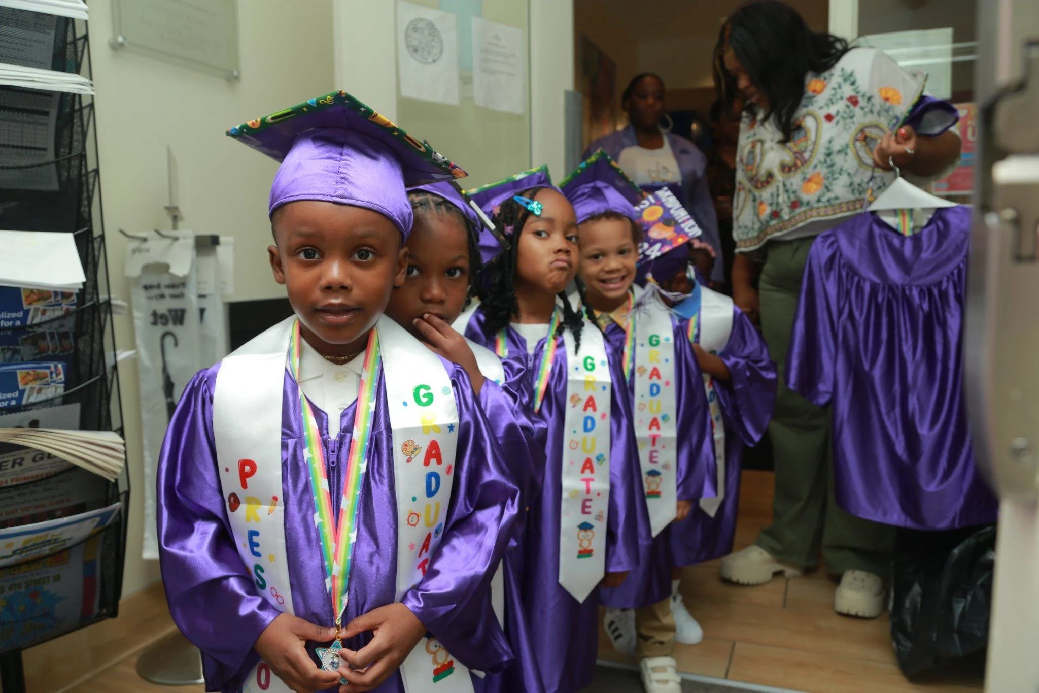 Line of young children wearing purple graduation gowns and hats, decorated with colorful medals and scarves, standing inside a room with an adult woman in the background, preparing for a graduation ceremony.