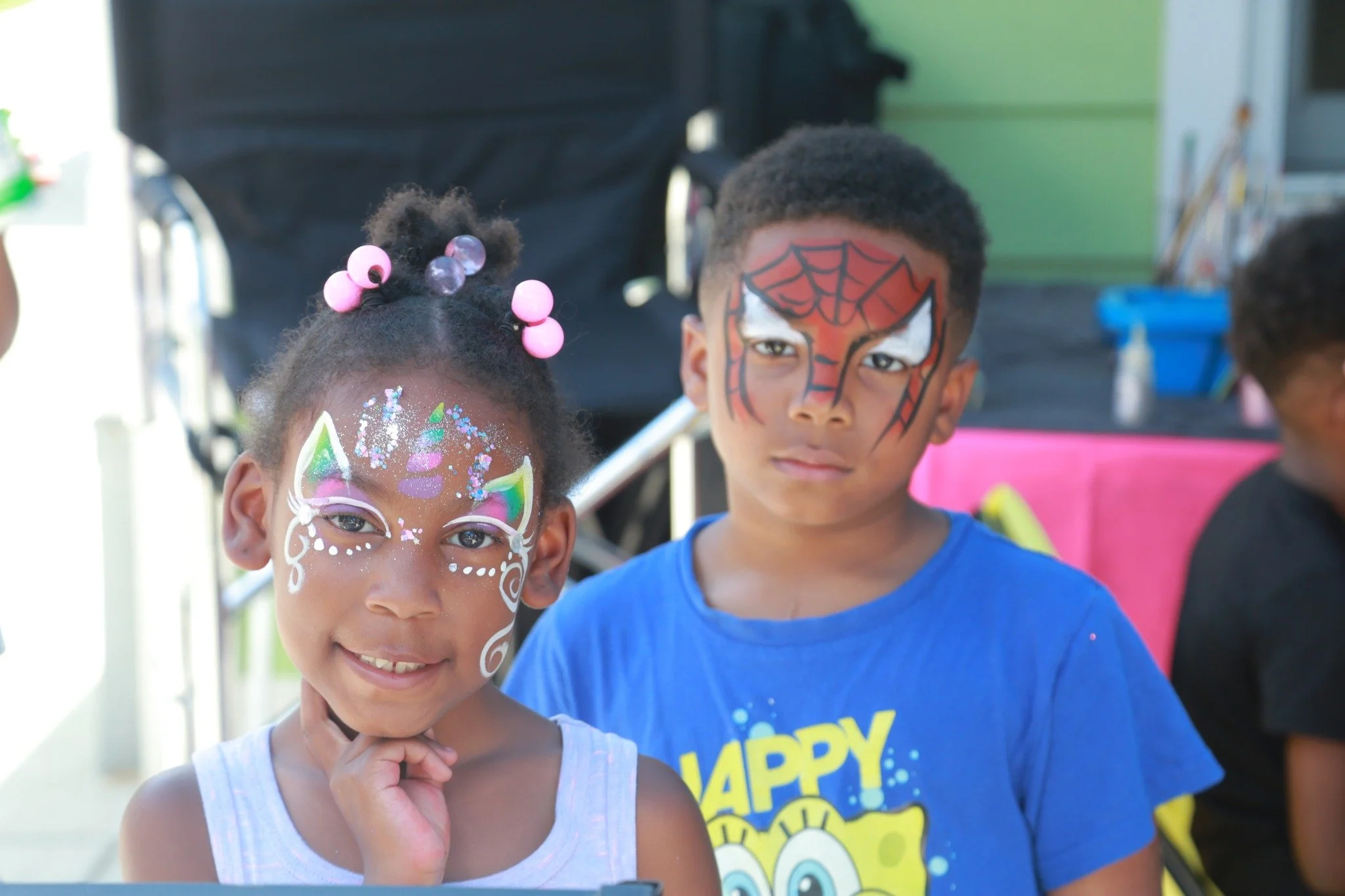Two kids with face paint at a celebration; a girl with a unicorn face design with glitter and beads, and a boy with Spider-Man face paint on his face.