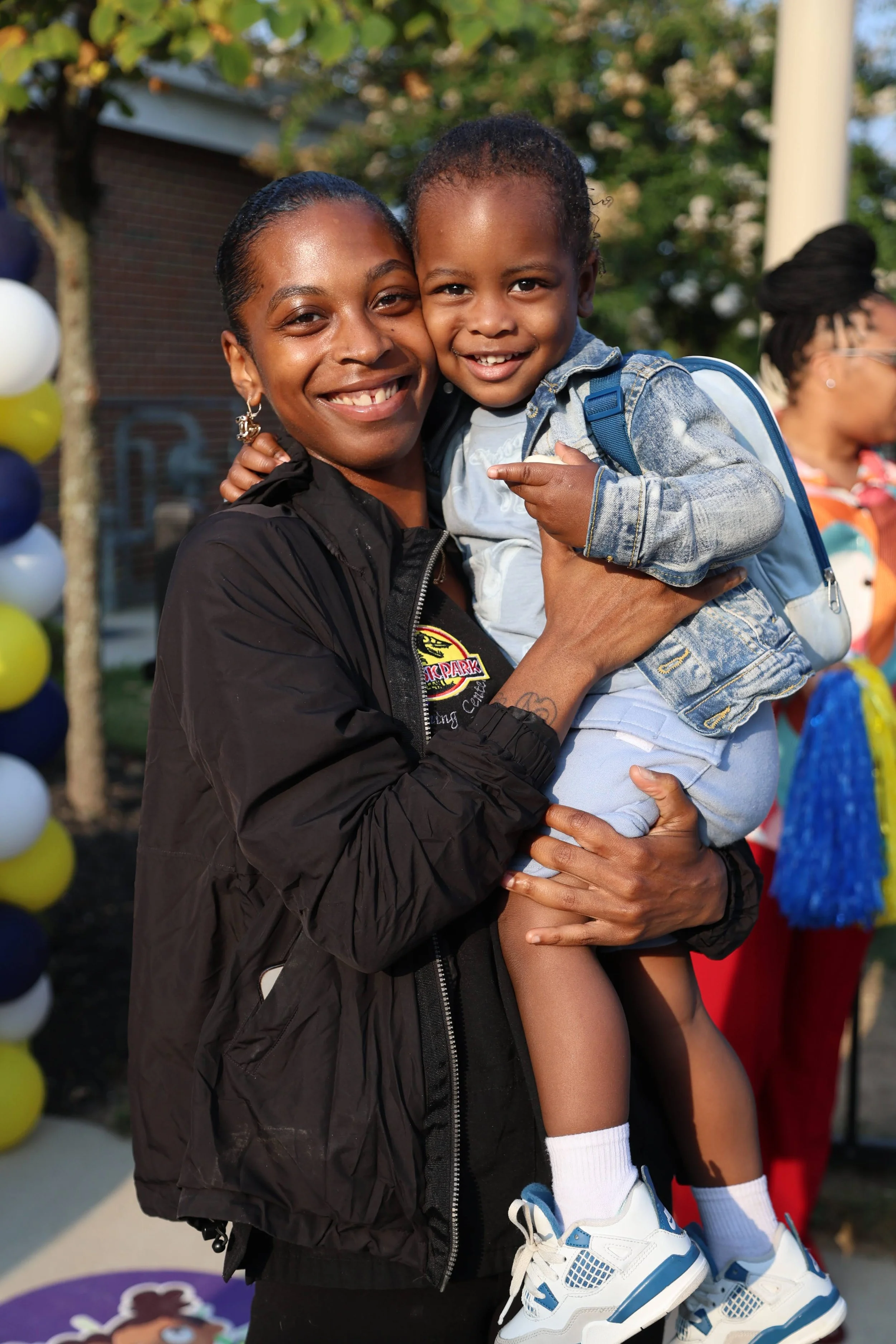 A woman smiling and holding a young boy, both appearing happy outdoors with balloons in the background.