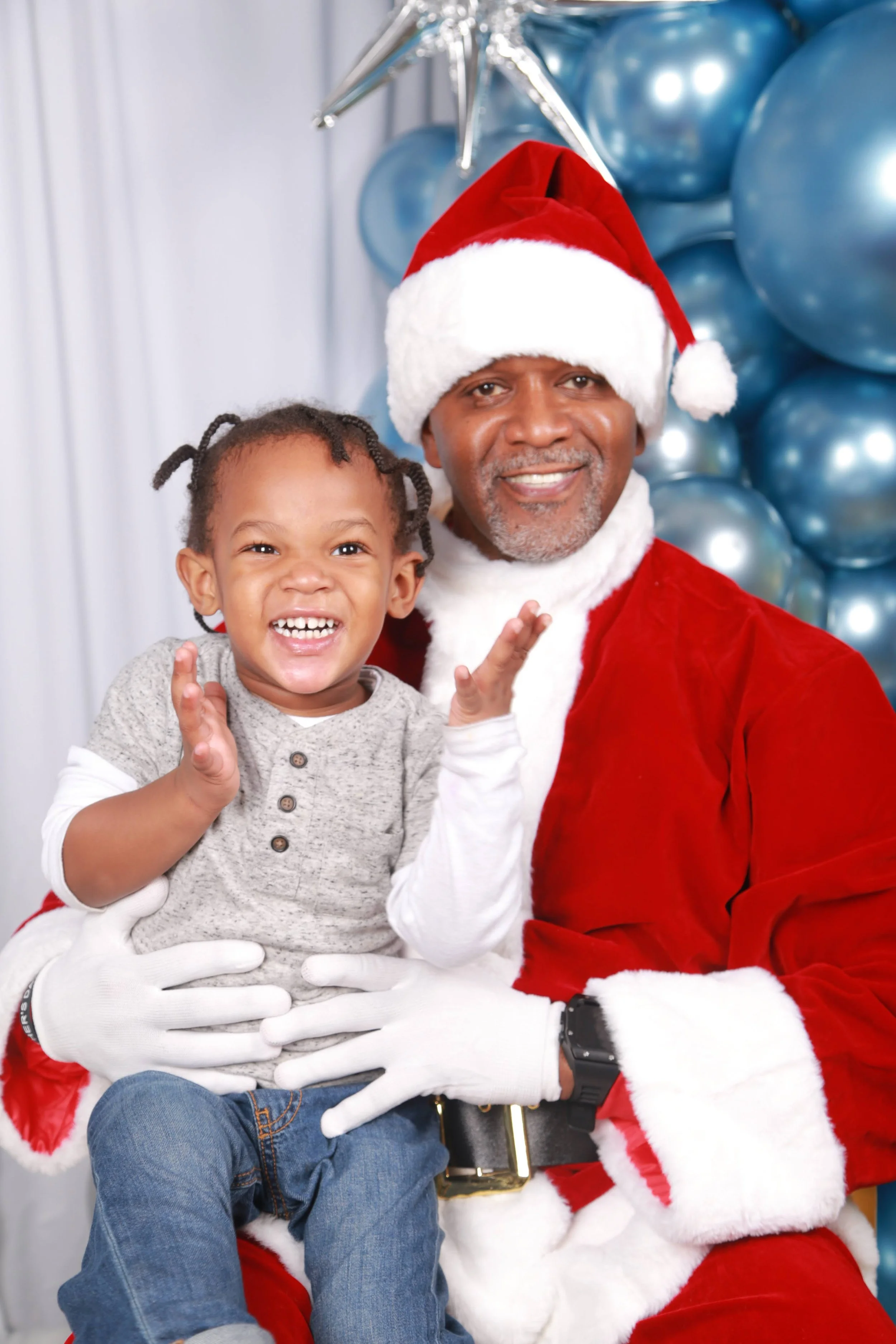 A young child sitting on Santa Claus's lap, both smiling, with blue balloon decorations in the background.
