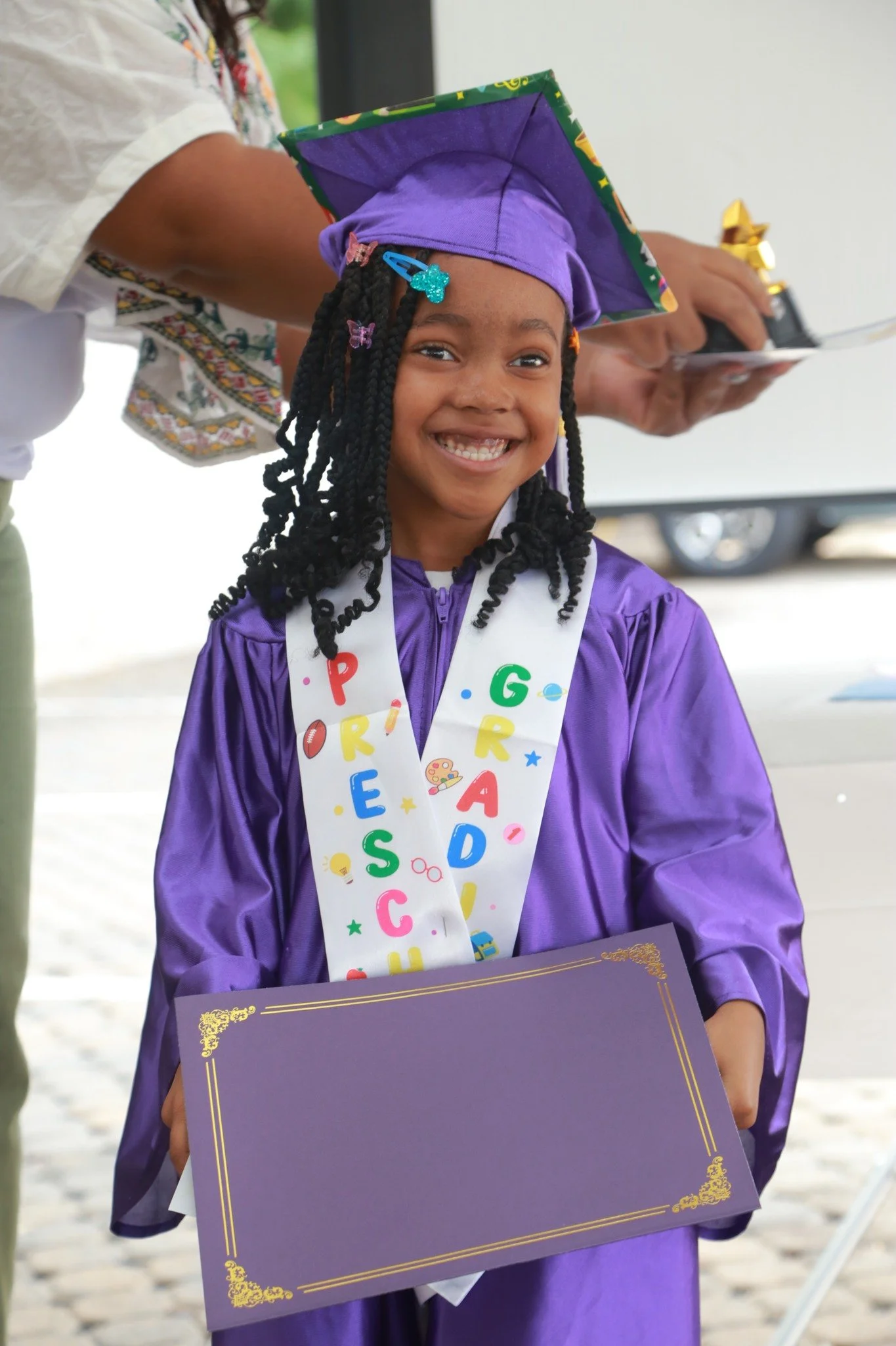 A young girl in a purple graduation gown and cap smiling while holding a blank purple certificate, wearing a white friendship cord with colorful letters and decorations, at a graduation ceremony.