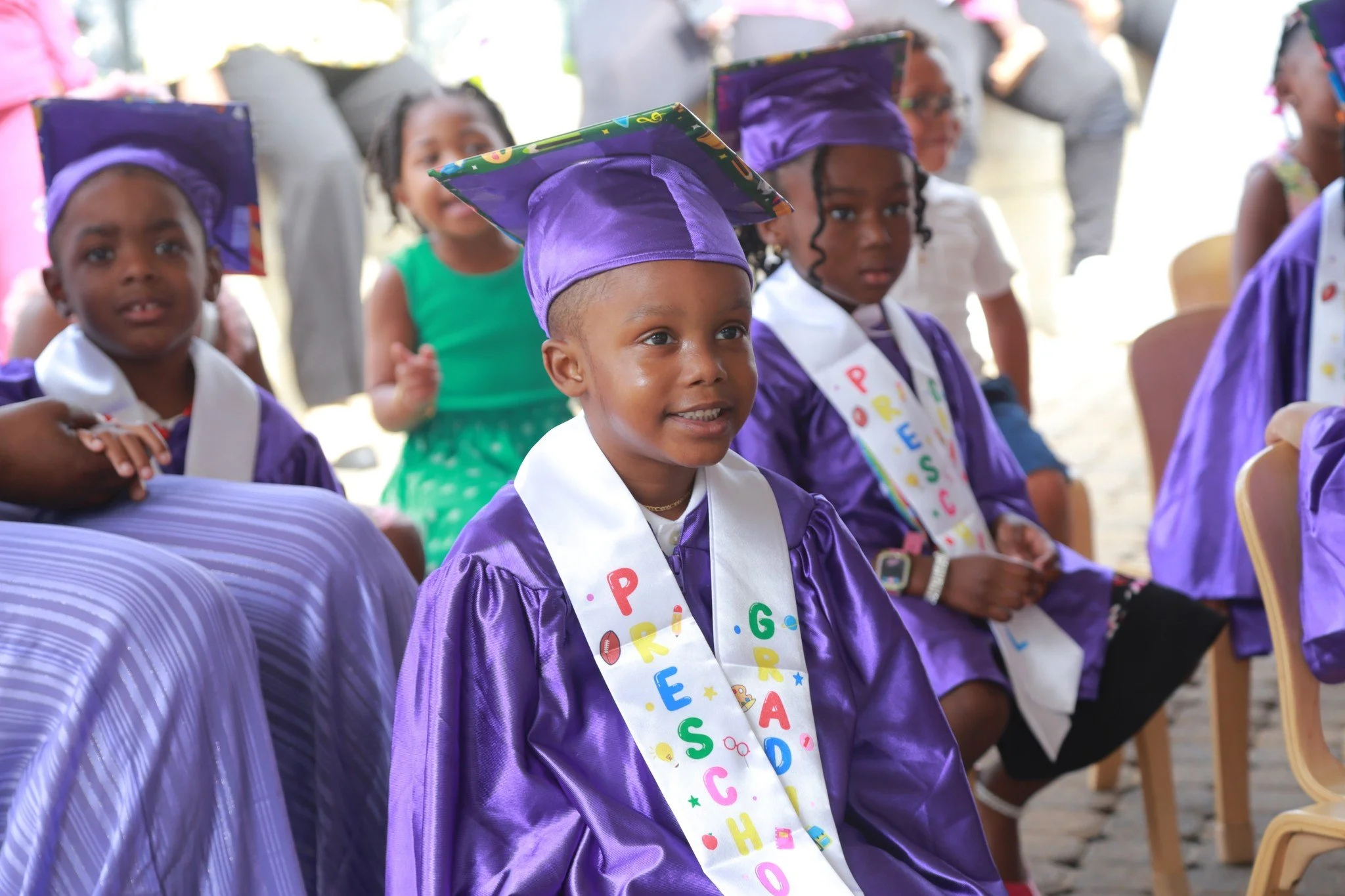 Young children wearing purple graduation gowns and caps, sitting and attending a graduation ceremony, with some wearing colorful sashes.