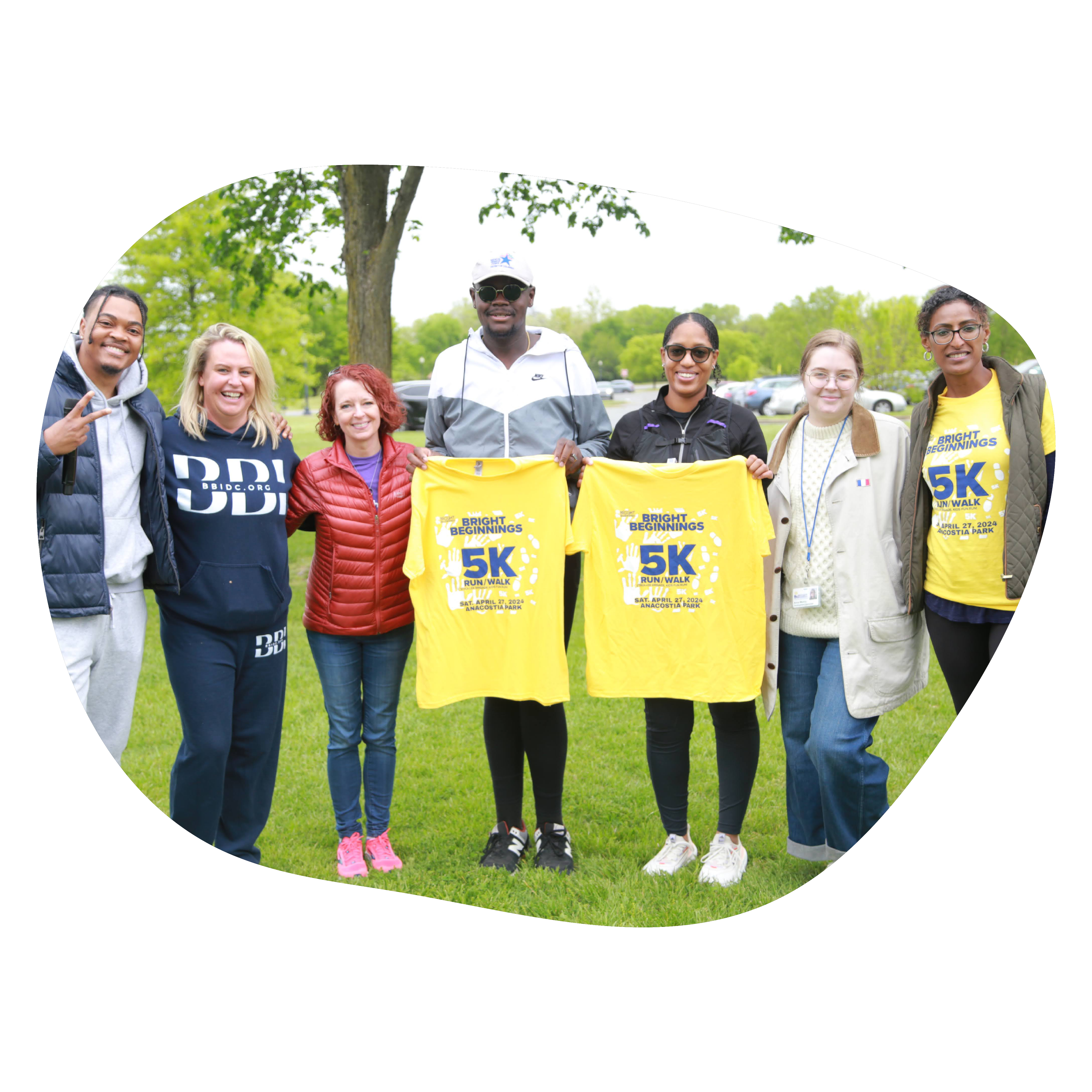 Group of seven diverse people standing outdoors at a park, holding yellow shirts with a 5K run/walk event logo, smiling, and dressed in casual clothing.
