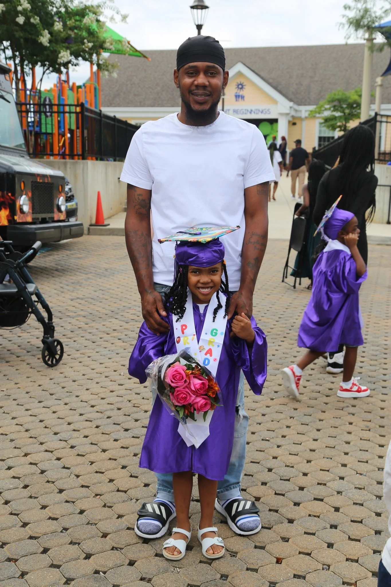 A young girl in a purple graduation gown and cap holding a bouquet of pink roses standing with an adult man in casual clothing, both smiling, at a graduation ceremony outdoors.