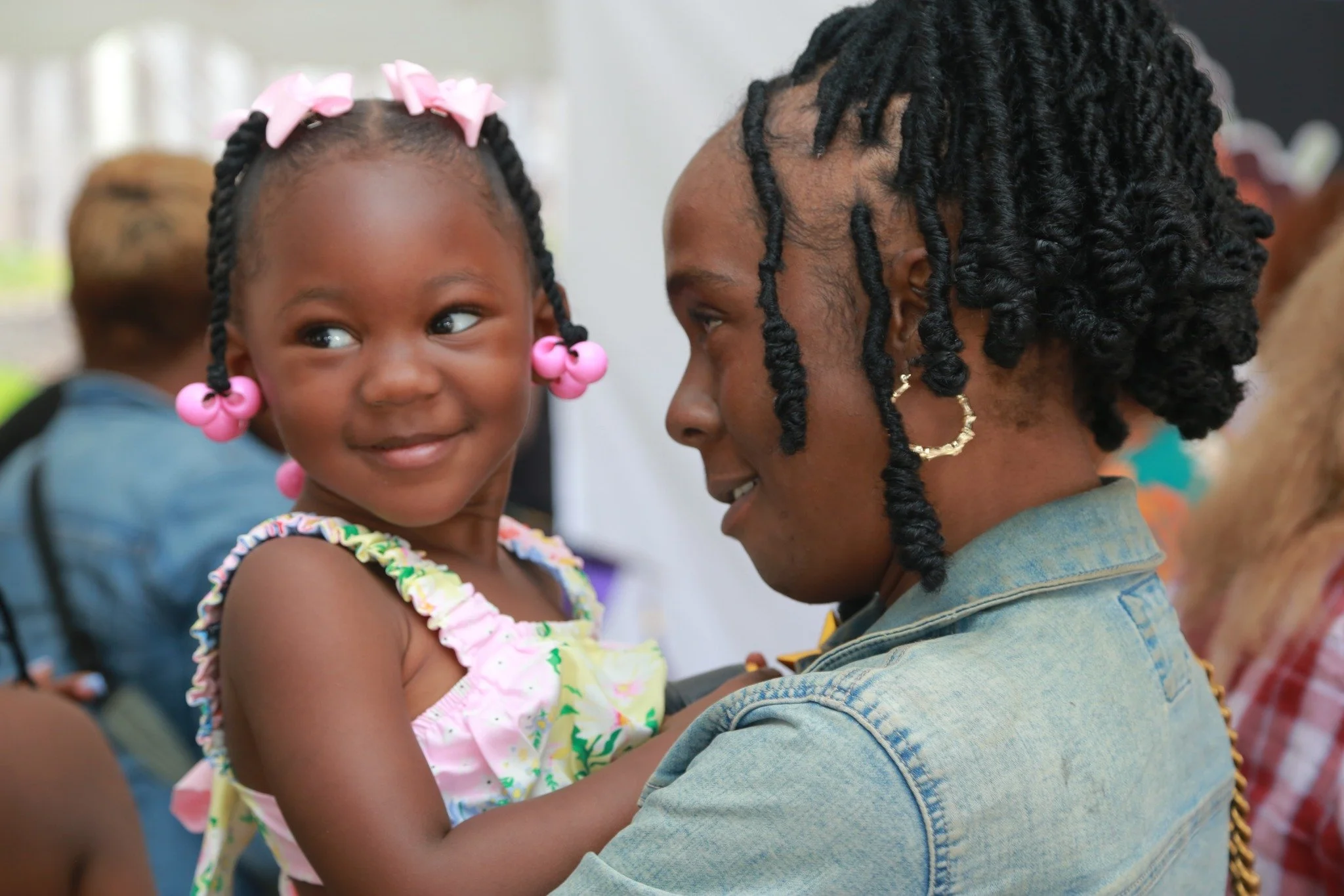 A young girl with pink bows in her braided hair, smiling while being held by a woman with styled dreadlocks, wearing hoop earrings, in a casual outdoor setting.