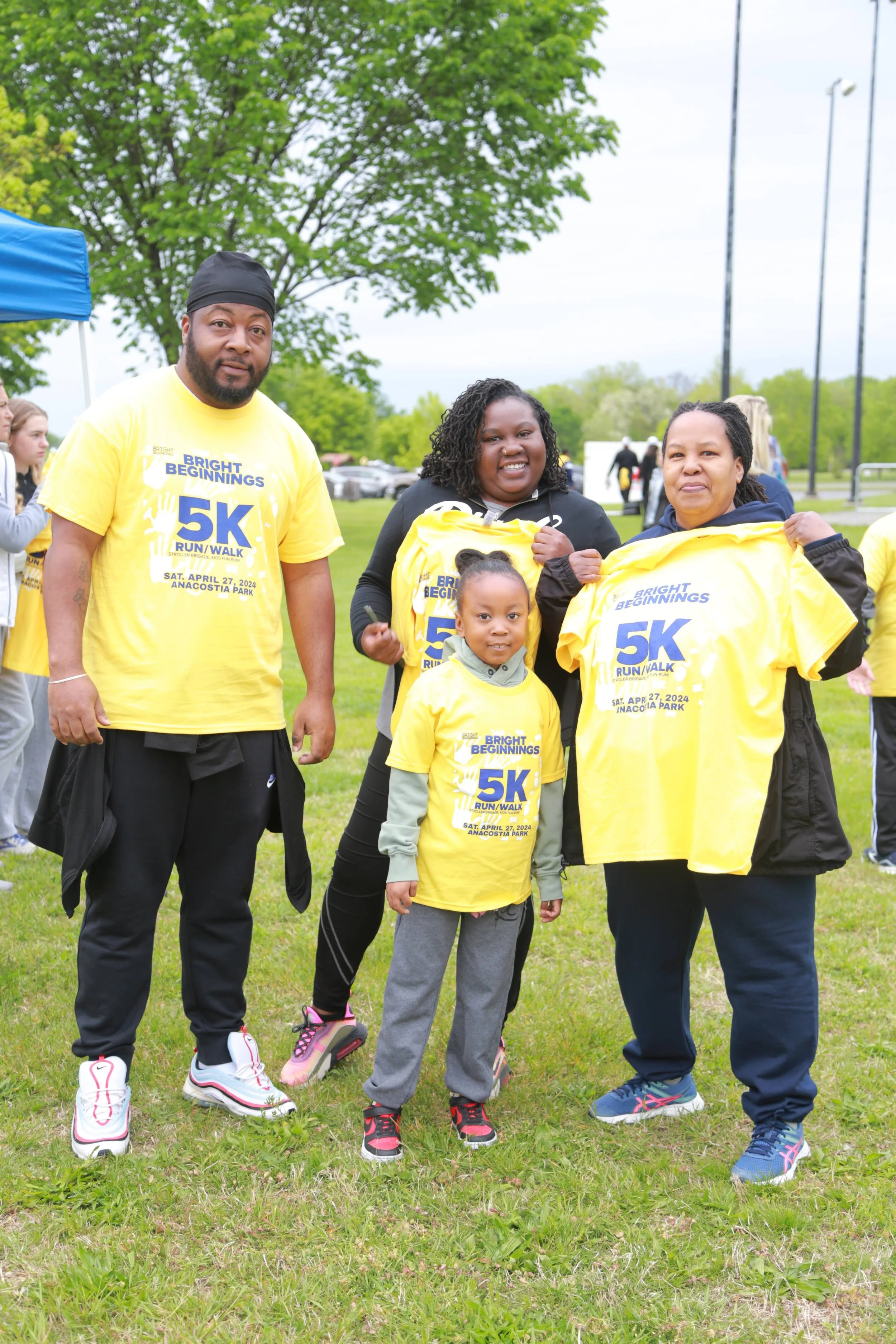 A group of four people, two adults and two children, standing outdoors at a park, wearing yellow T-shirts for a 5K run/walk event. They are smiling, holding or displaying their event shirts. The background shows trees, other event participants, and a