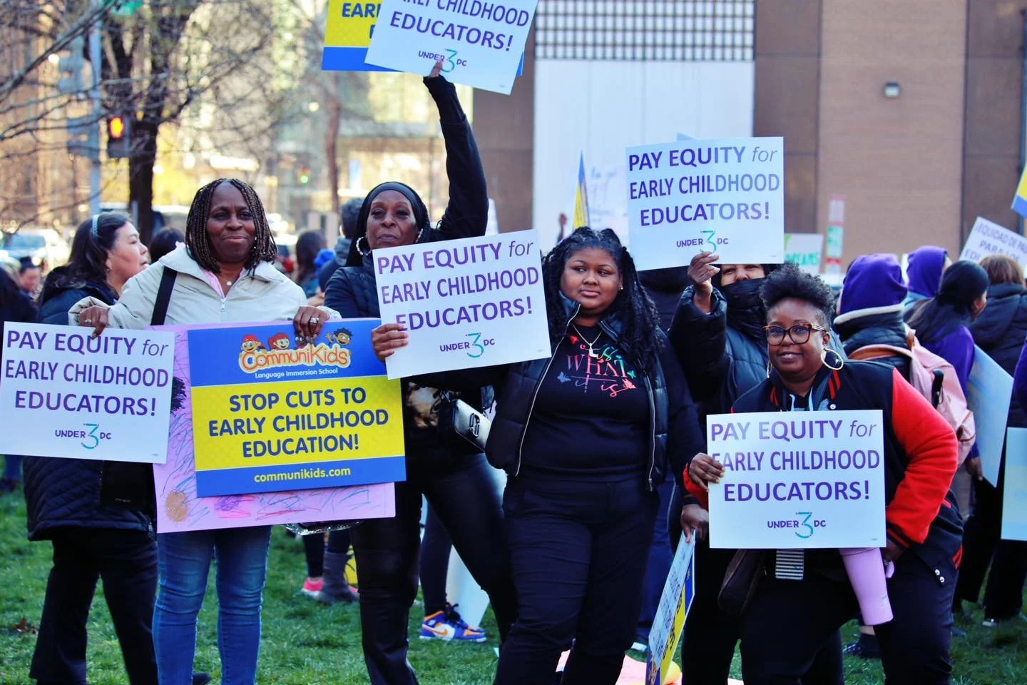A diverse group of women holding signs that advocate for paying early childhood educators and stopping cuts to early childhood education during a protest.