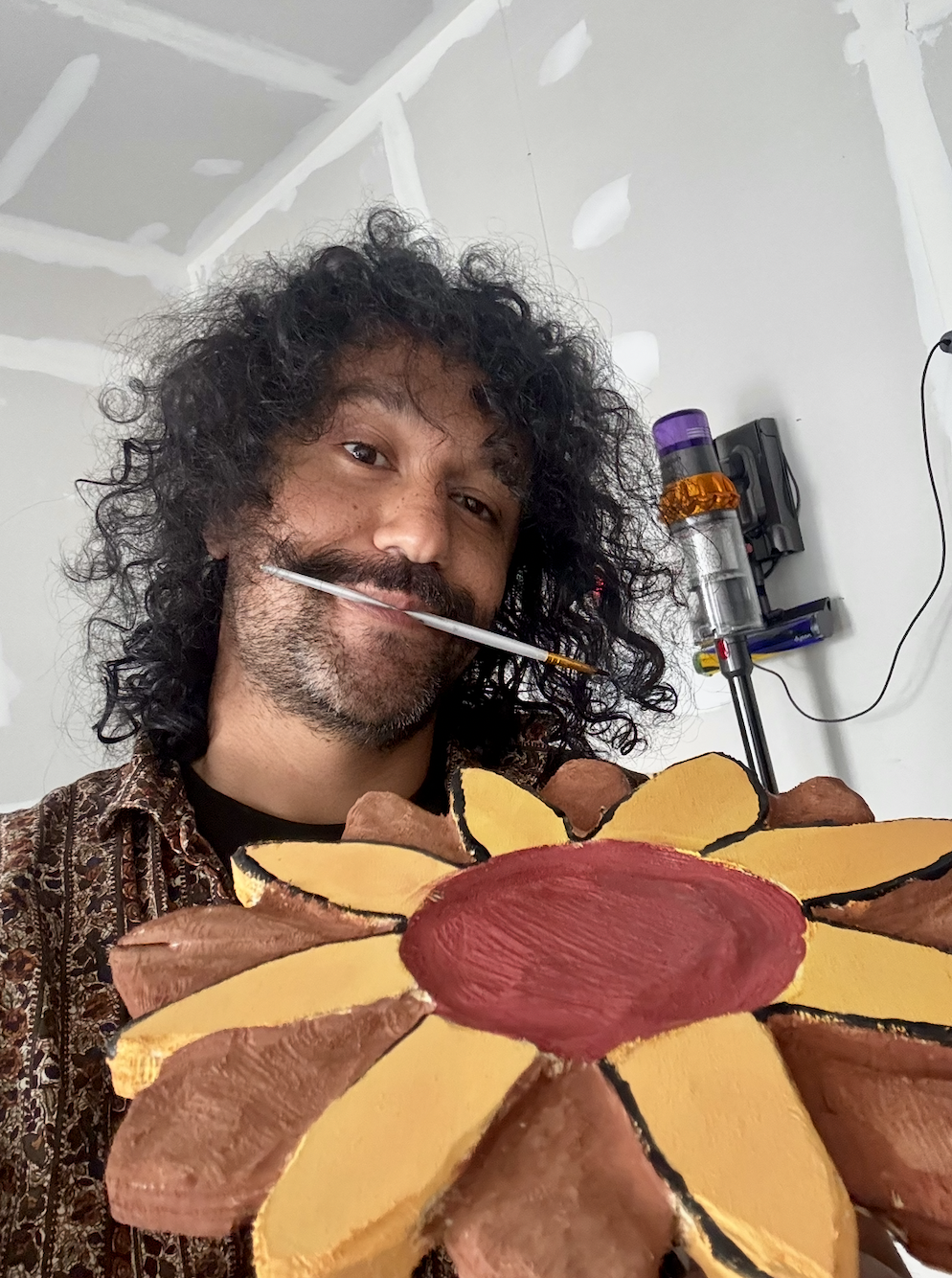 Sawtistic with curly black hair and a beard is holding a colorful sunflower-shaped sculpture. He has a yellow and brown painted face. He is smiling and has a paintbrush in his mouth. He is digging his Bob Ross look