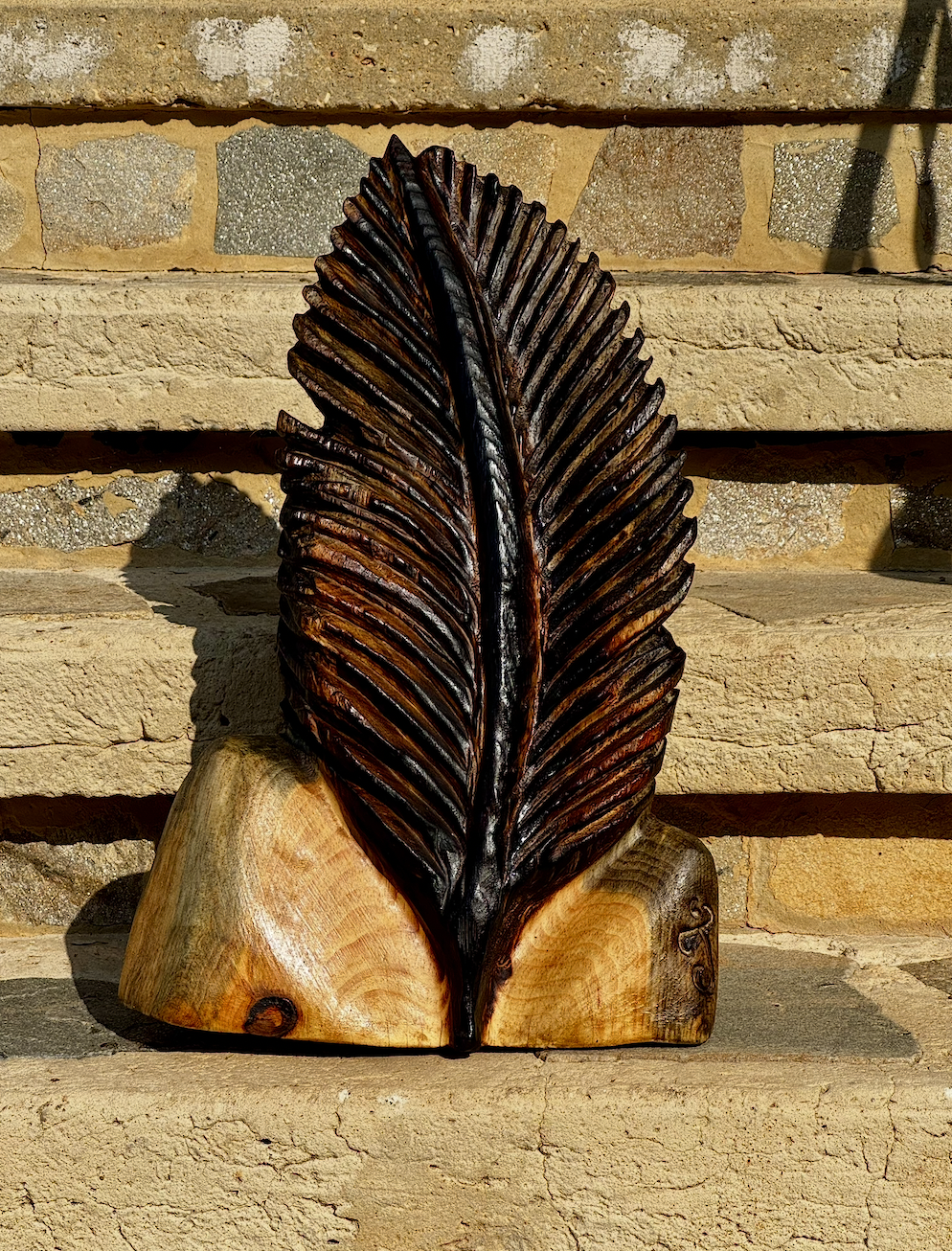 A carved wooden art piece resembling a large leaf, mounted on a wooden base, with a brick wall background.