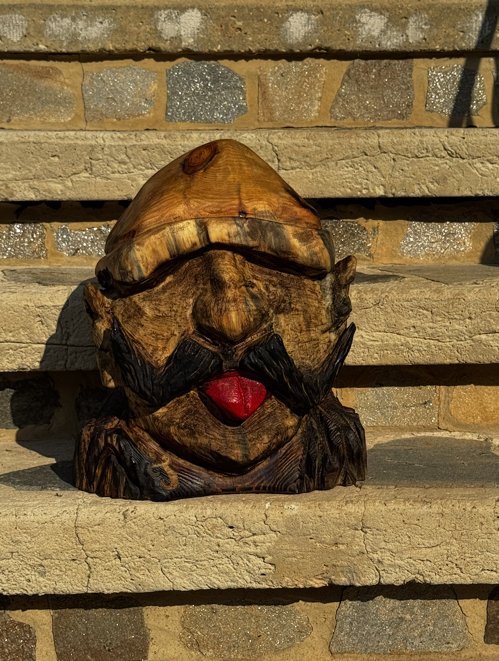 Wooden sculpture of a face with a red tongue and black beard, placed on a concrete step in front of a brick wall.