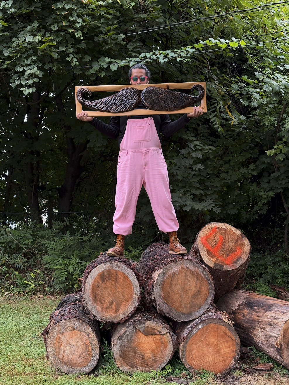 Sawtistic standing on a pile of cut logs outdoors, holding a large wooden frame with a carved wooden mustache, wearing pink overalls, black shirt, sunglasses, and brown boots, with green foliage in the background.
